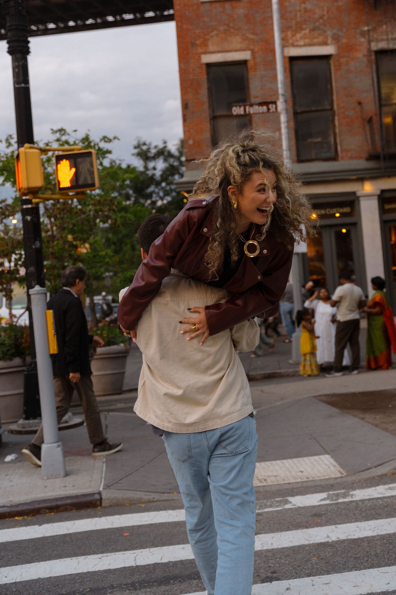 A person in jeans and a brown jacket dances playfully on a city sidewalk near brick buildings and pedestrians.