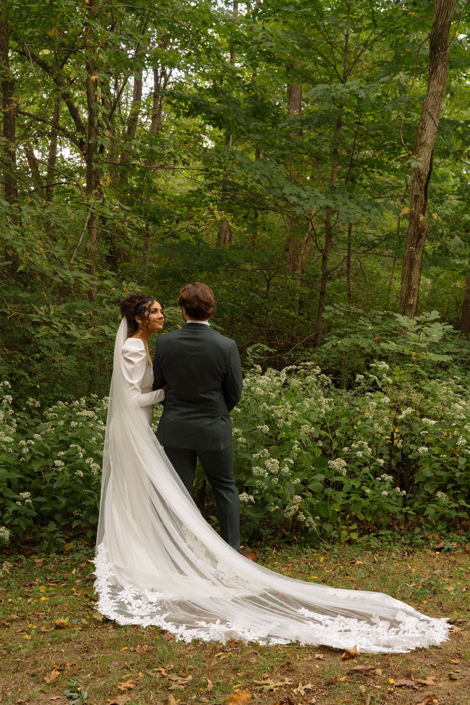 Bride and groom walk through wooded path with long flowing cathedral veil trailing behind.