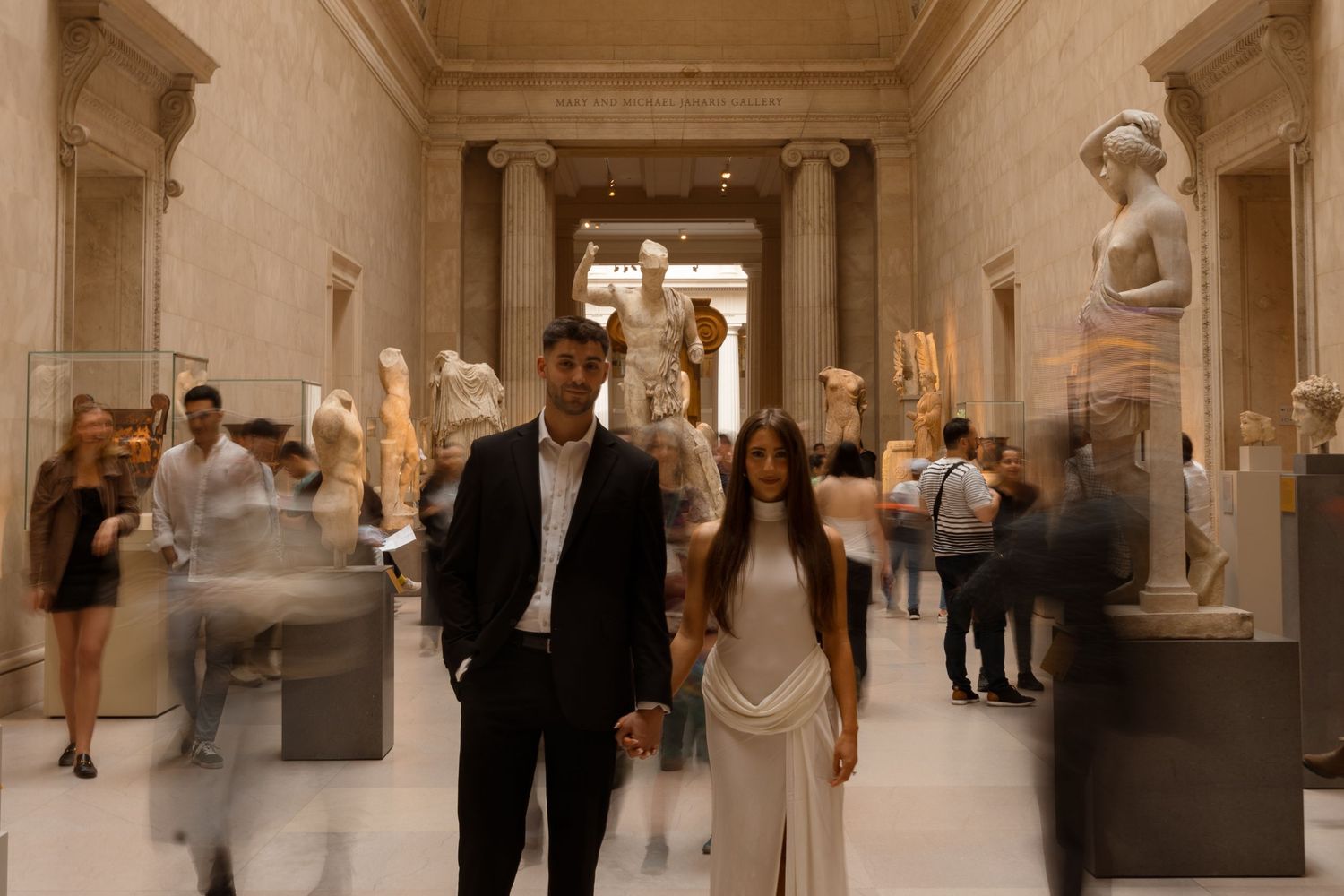 A couple walks through an elegant museum gallery filled with classical statues and artwork.