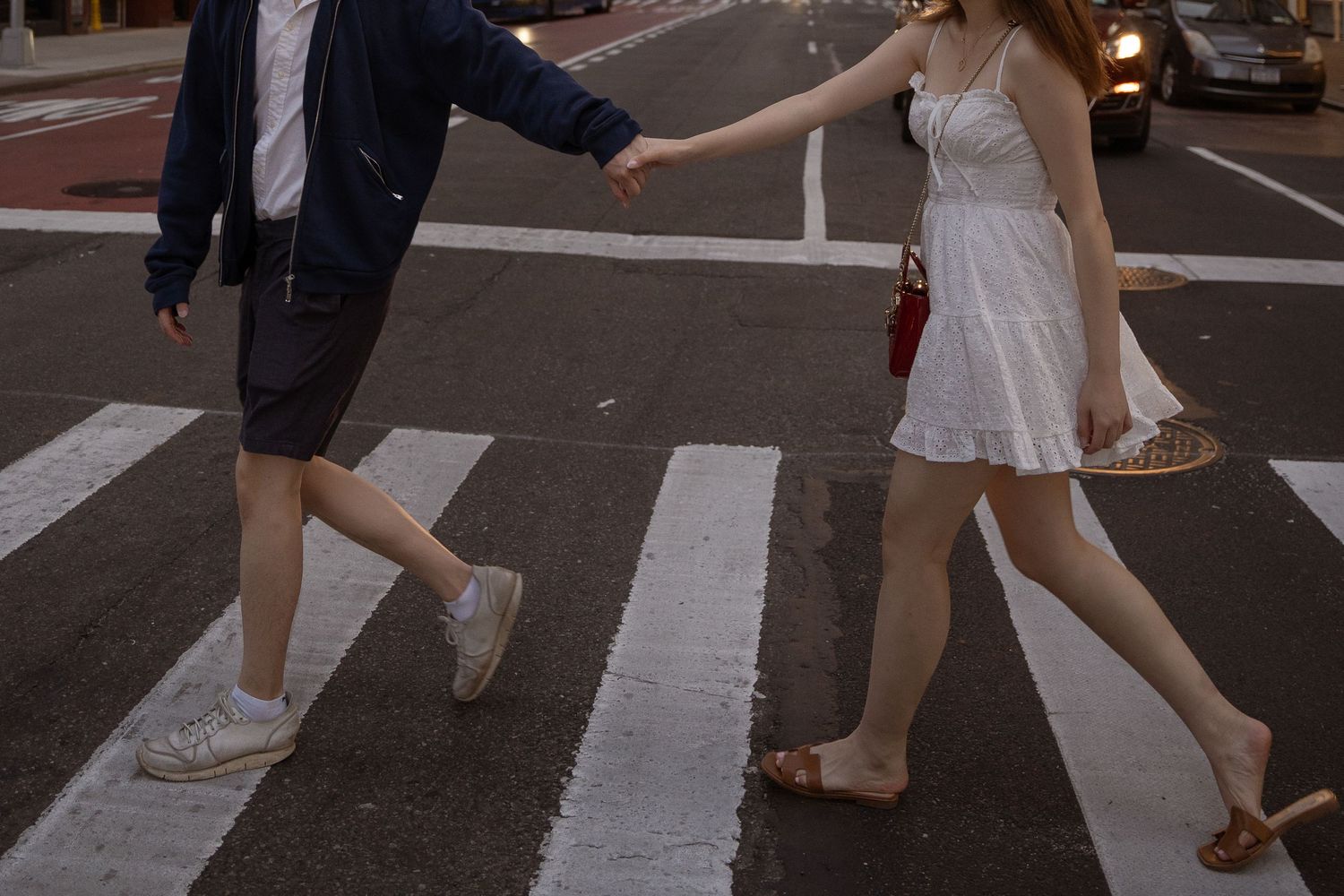 Couple crossing a city street crosswalk at dusk with urban buildings in background.