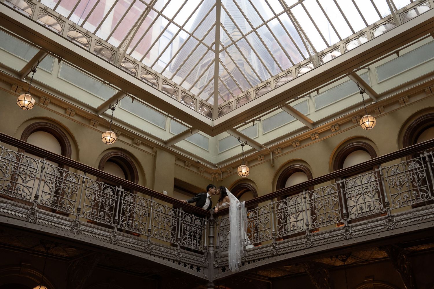 Elegant glass ceiling architecture with ornate railings and hanging pendant lights in a wedding venue.