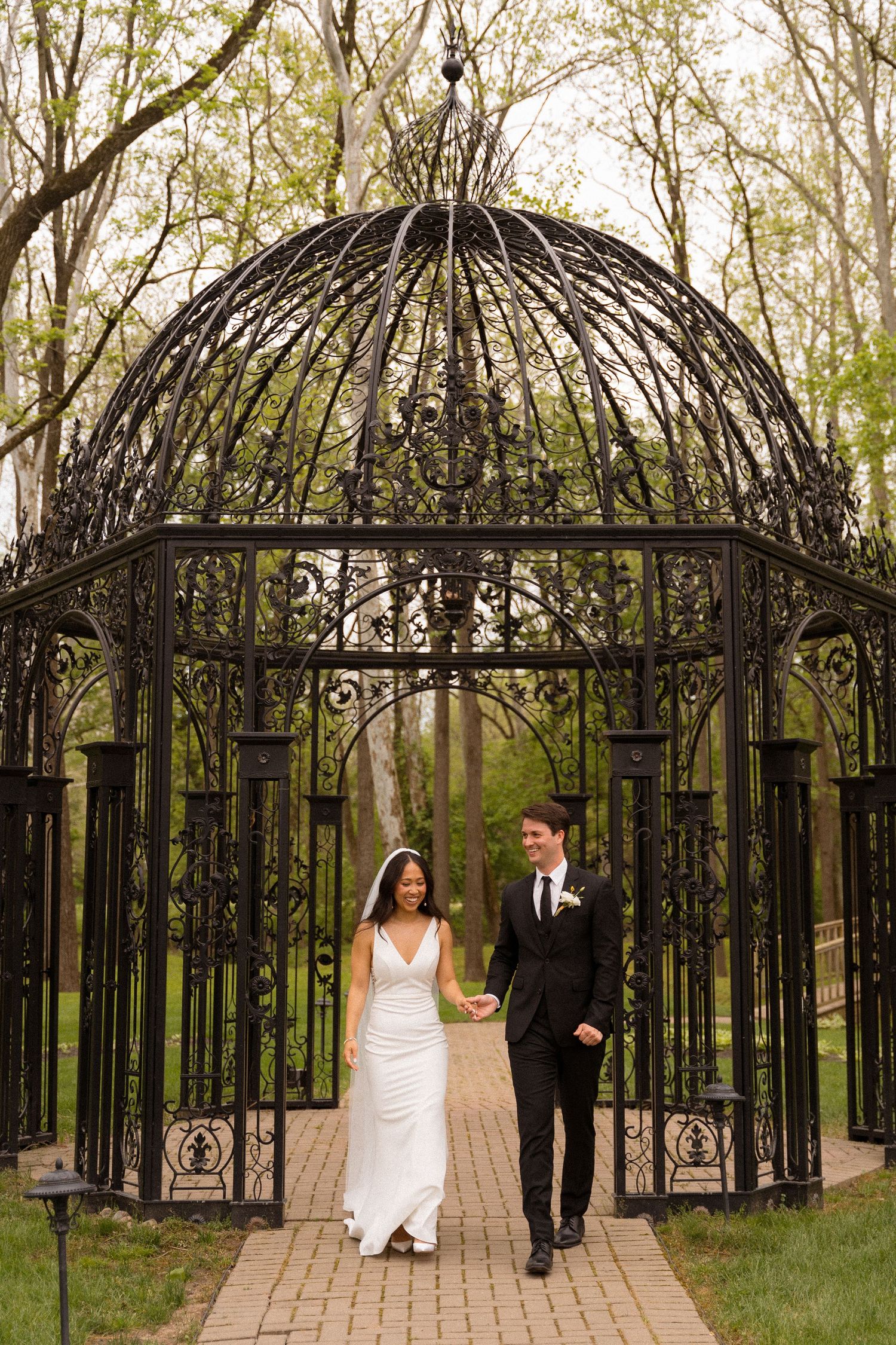 A couple walks beneath an ornate wrought iron garden gazebo with a domed top along a brick pathway in a wooded setting.