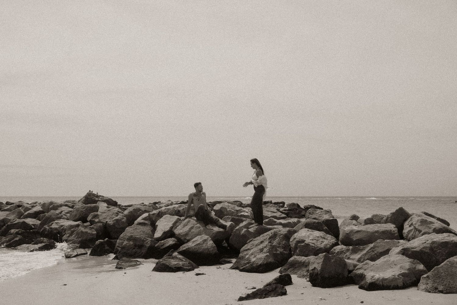 Two people standing on rocky jetty overlooking ocean in black and white.
