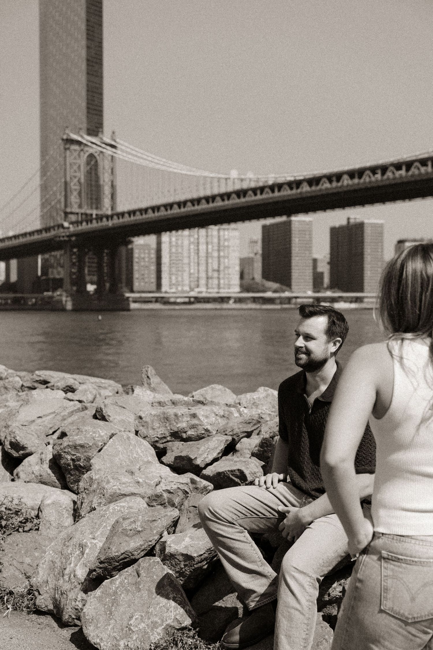 A couple relaxes on rocky shoreline with Manhattan Bridge visible in background.