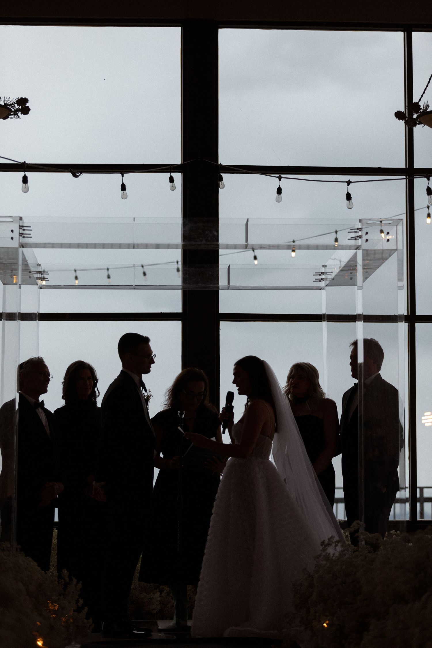 Silhouettes of wedding party members stand against window with hanging string lights and cloudy sky backdrop.