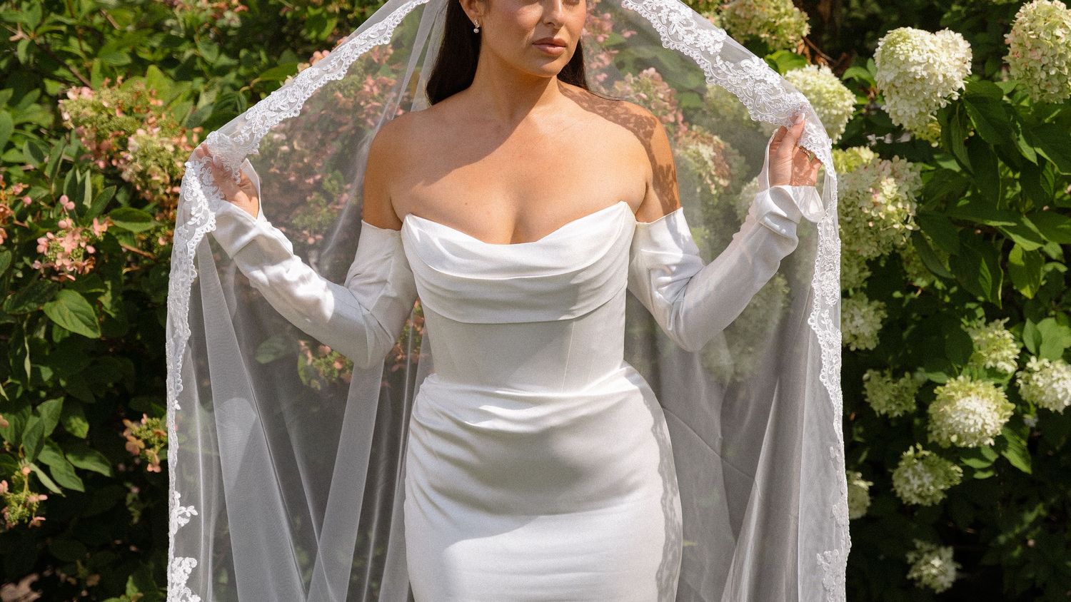 Elegant white wedding dress with draped neckline and flowing veil against floral garden backdrop.