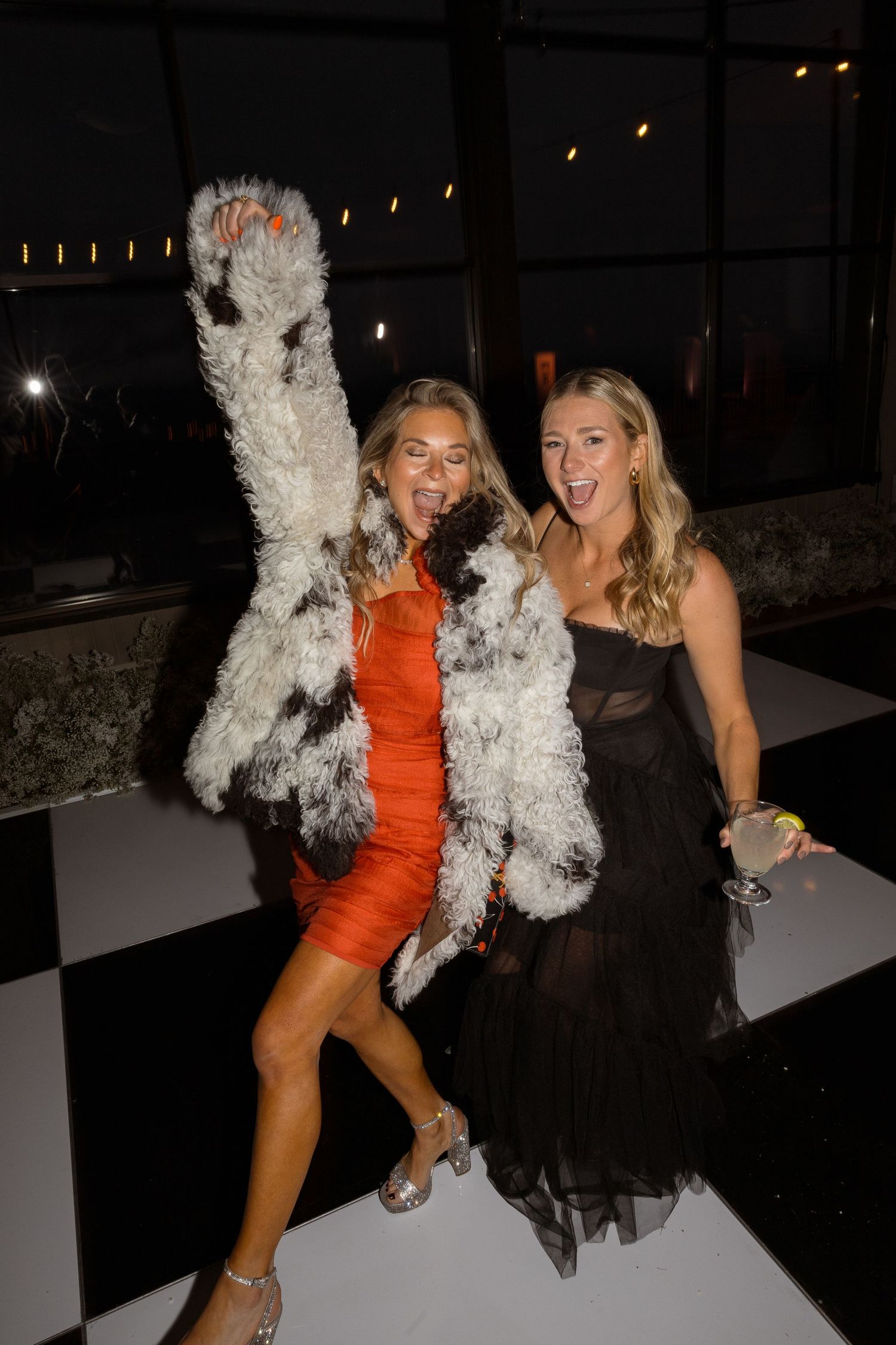 Two women in evening wear playfully pose with white feather boas at a nighttime event.
