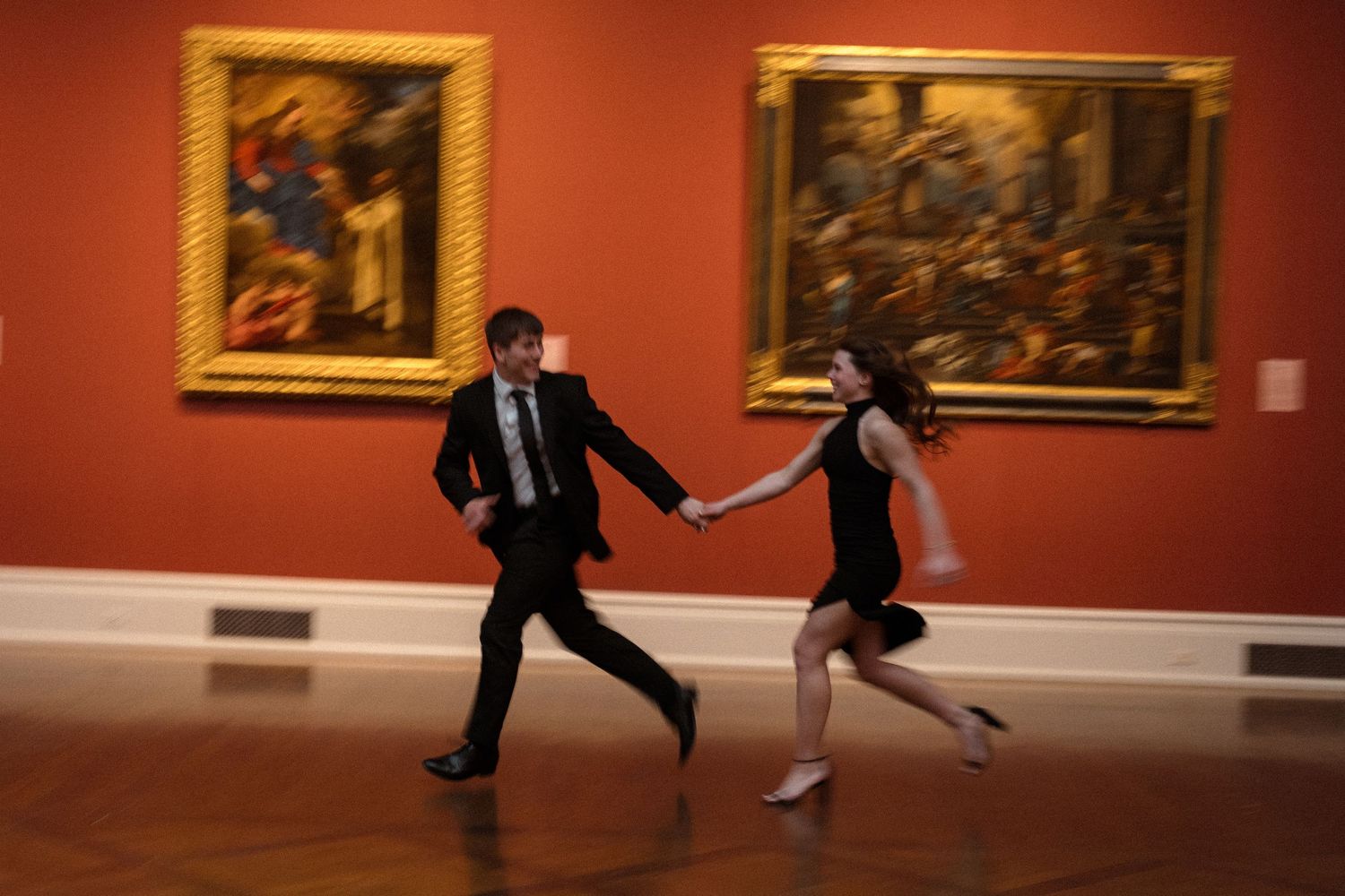 Two figures dance playfully in an art gallery with red walls and framed paintings.