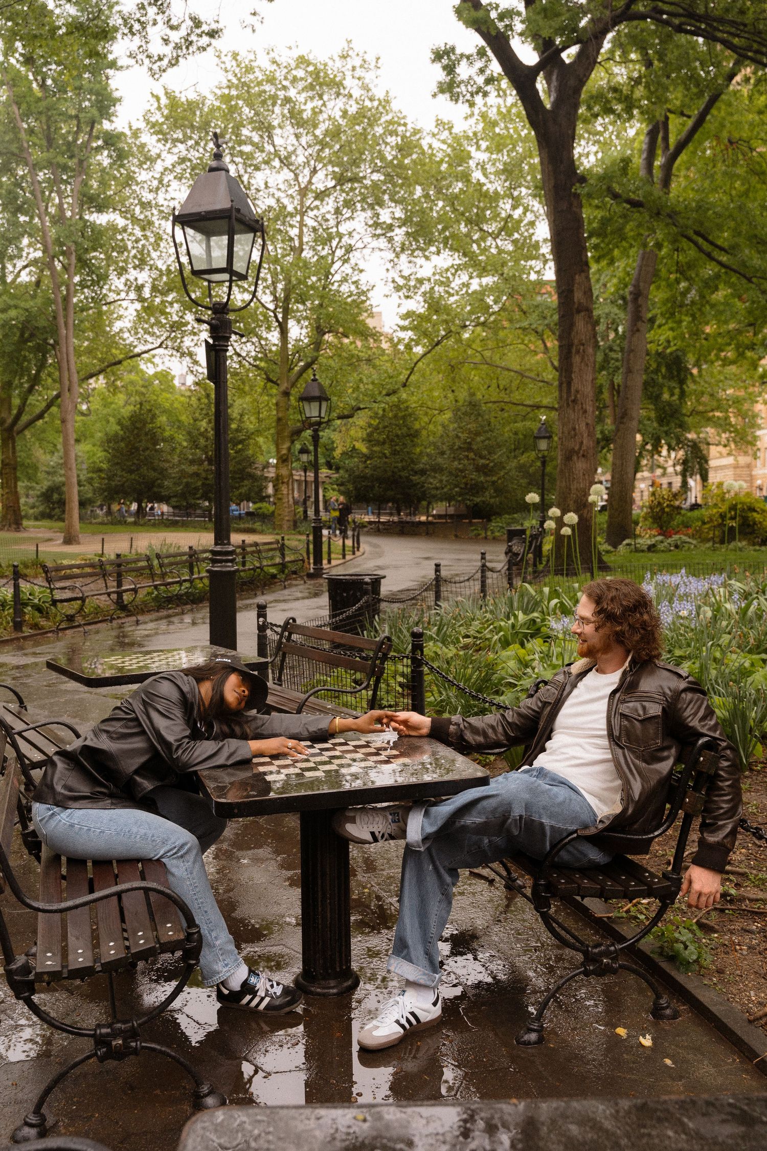 Two people play chess at a table in a park with vintage lampposts.
