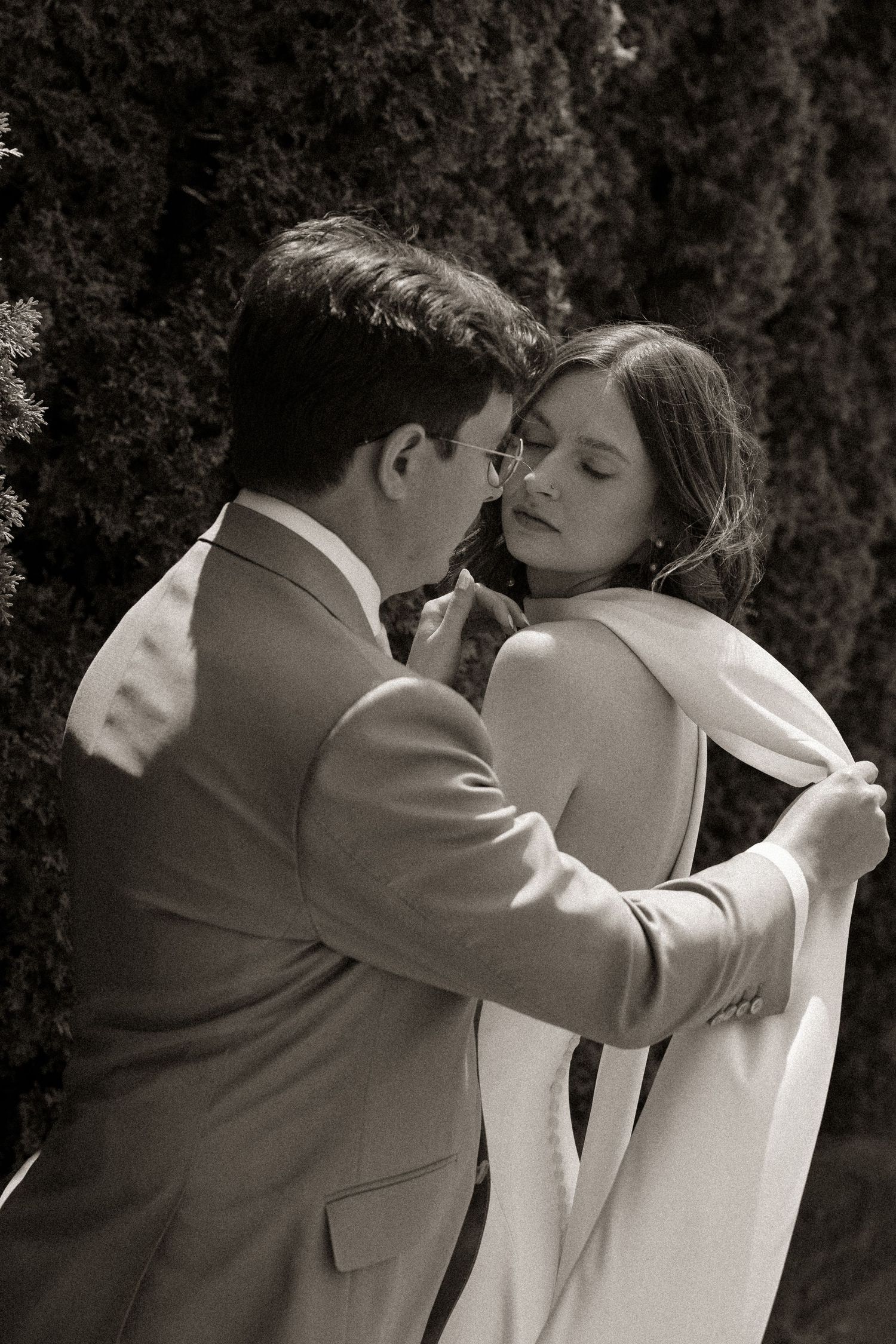 Intimate black and white portrait of couple embracing against dark foliage.