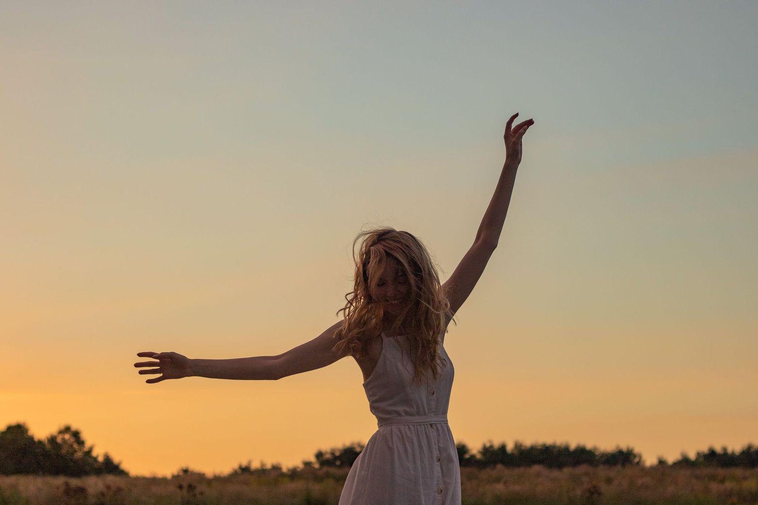 Person in white dress raising arms joyfully against a pastel sunset sky.