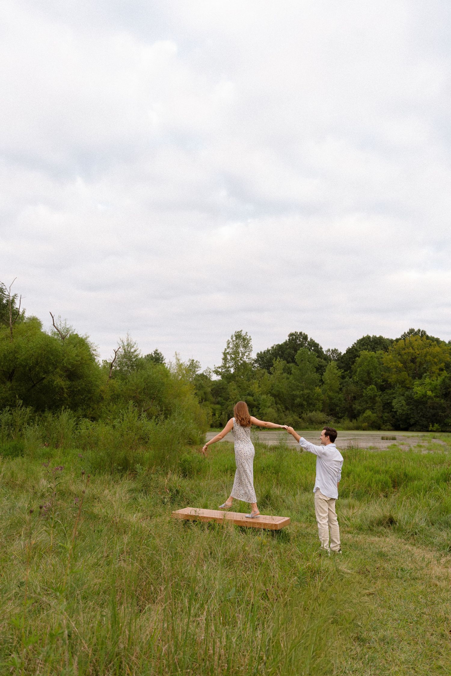 A couple balances on a wooden beam in a grassy field under cloudy skies.