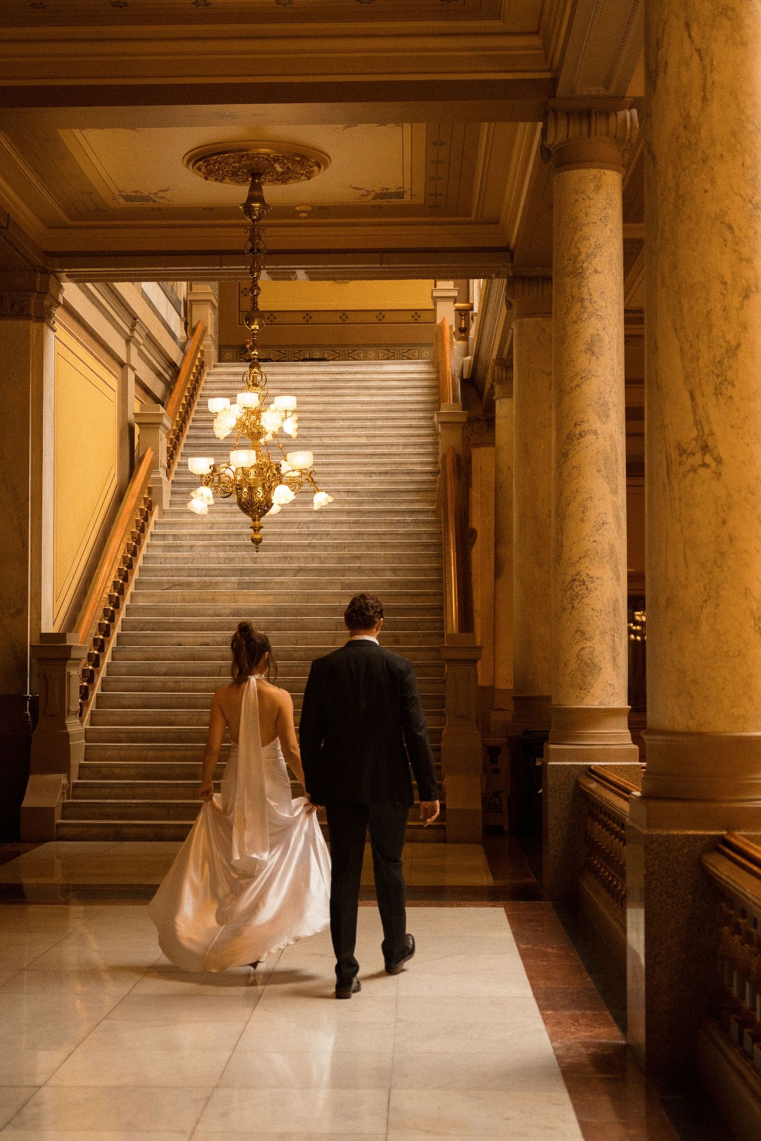 Elegant couple walking through a grand hallway with marble columns and chandeliers.