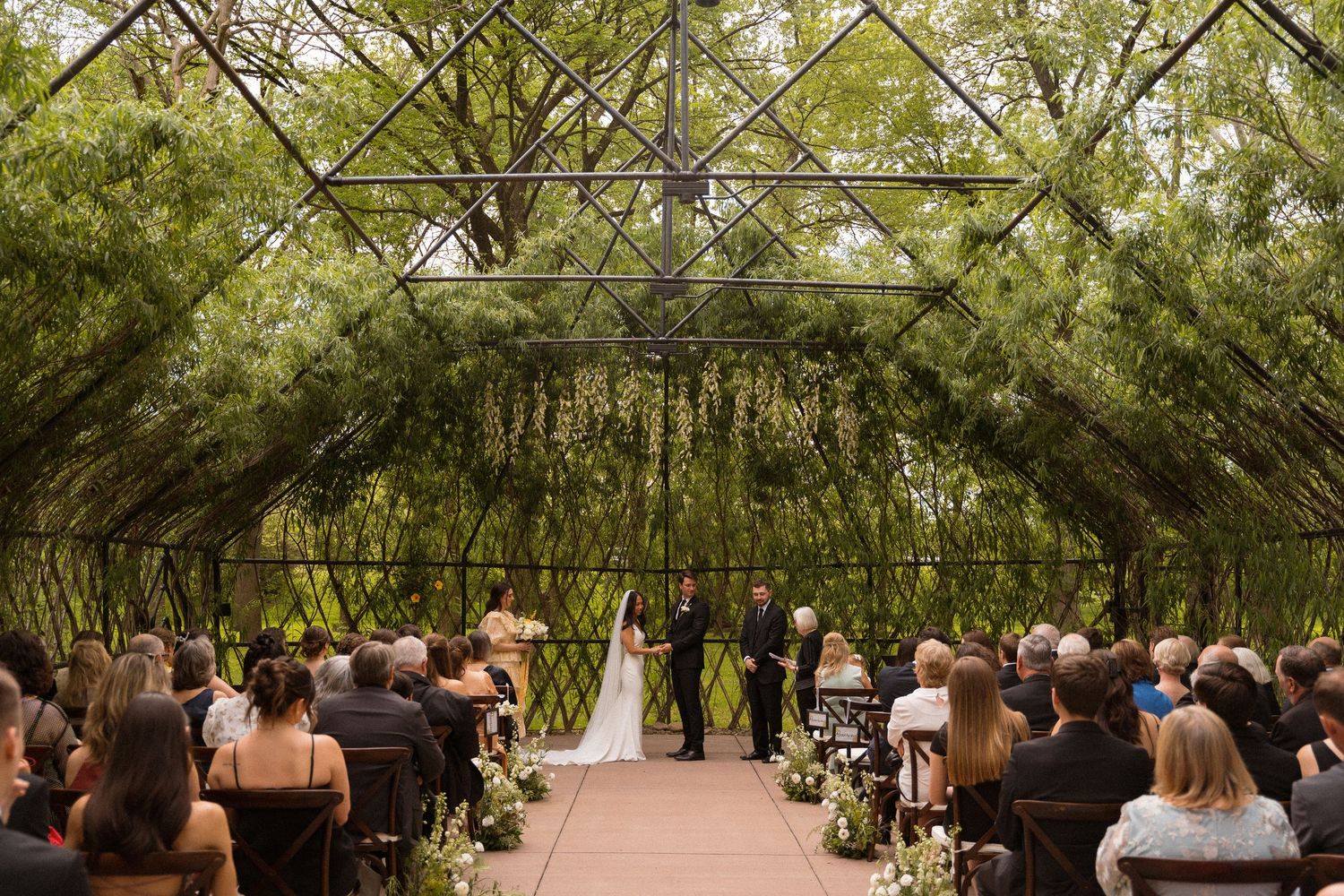 An outdoor wedding ceremony setup with wooden chairs under a natural green canopy.