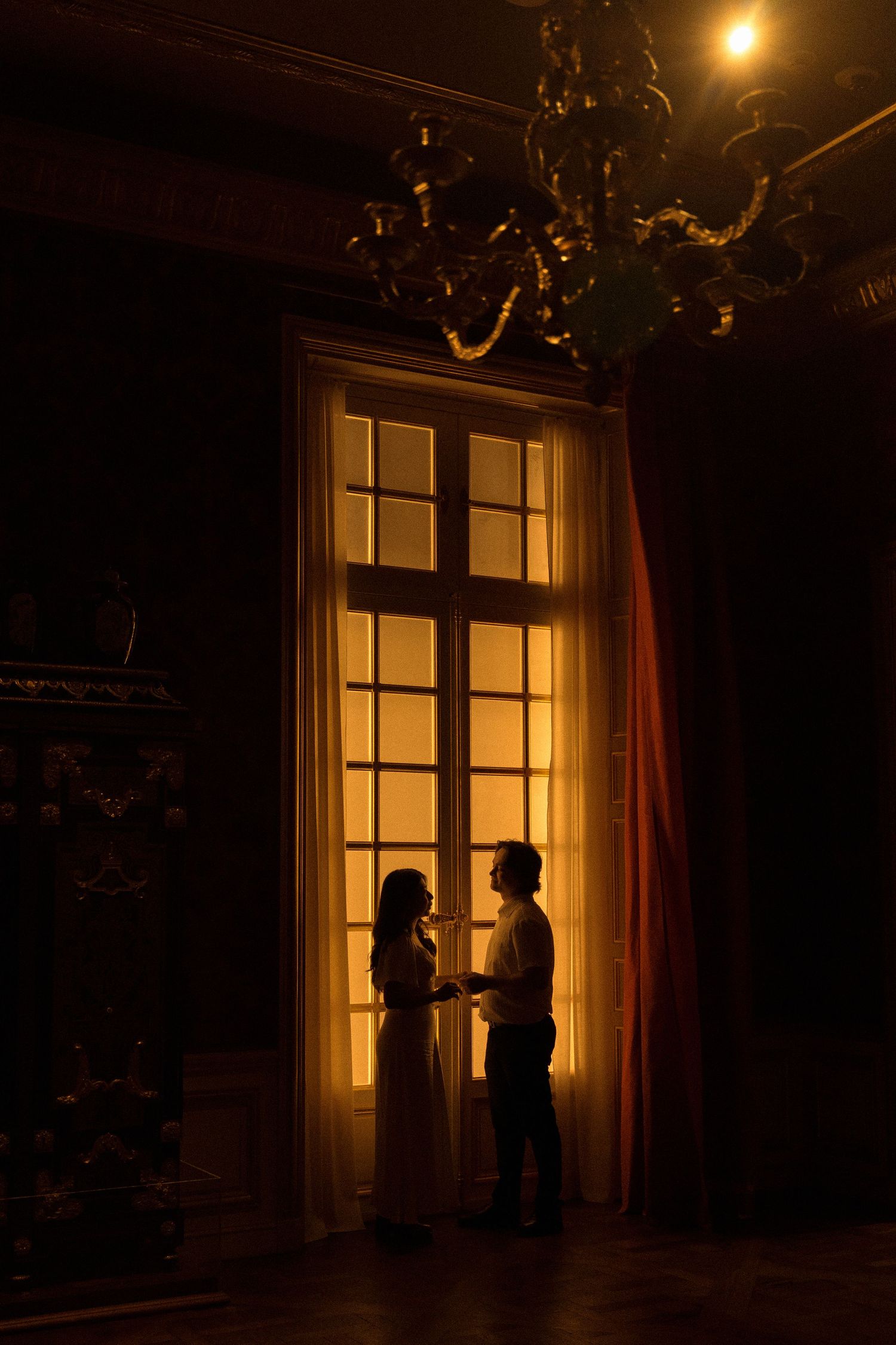 Silhouettes stand near illuminated windows in a dimly lit room creating a romantic atmosphere.