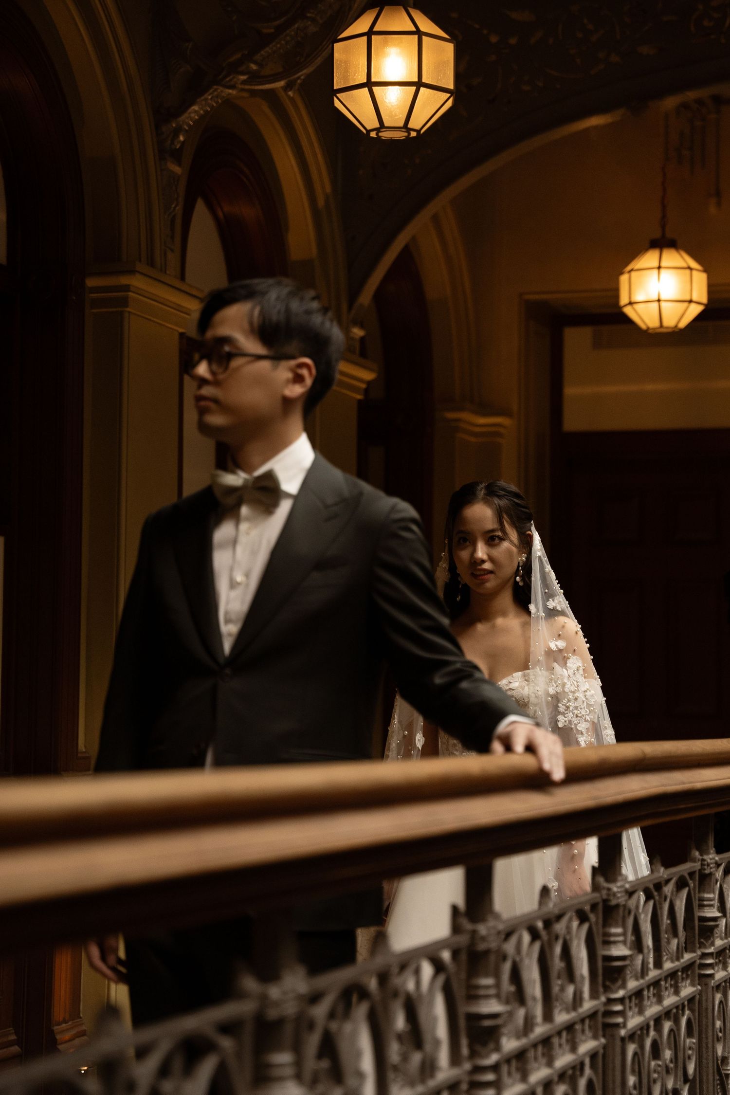 A couple in formal wedding attire stand together near an ornate railing with vintage lanterns overhead.
