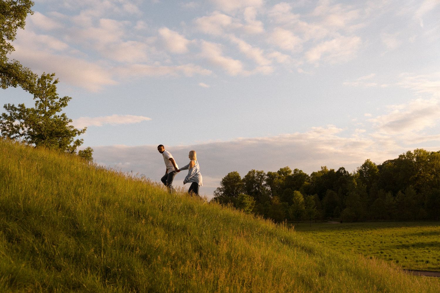 Two people walk hand in hand up a grassy hill during sunset.