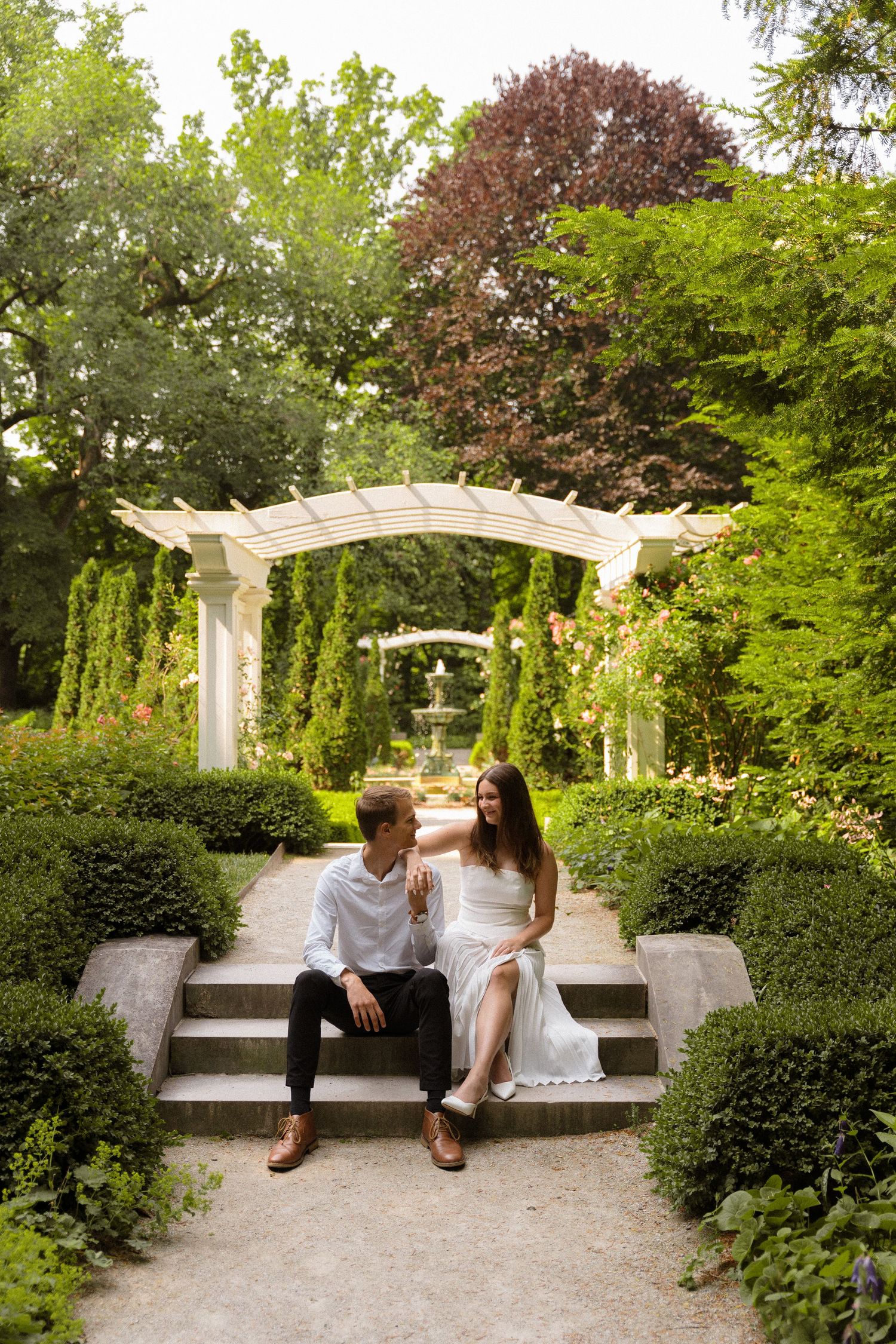 A couple sits on garden steps beneath a white pergola surrounded by lush greenery.