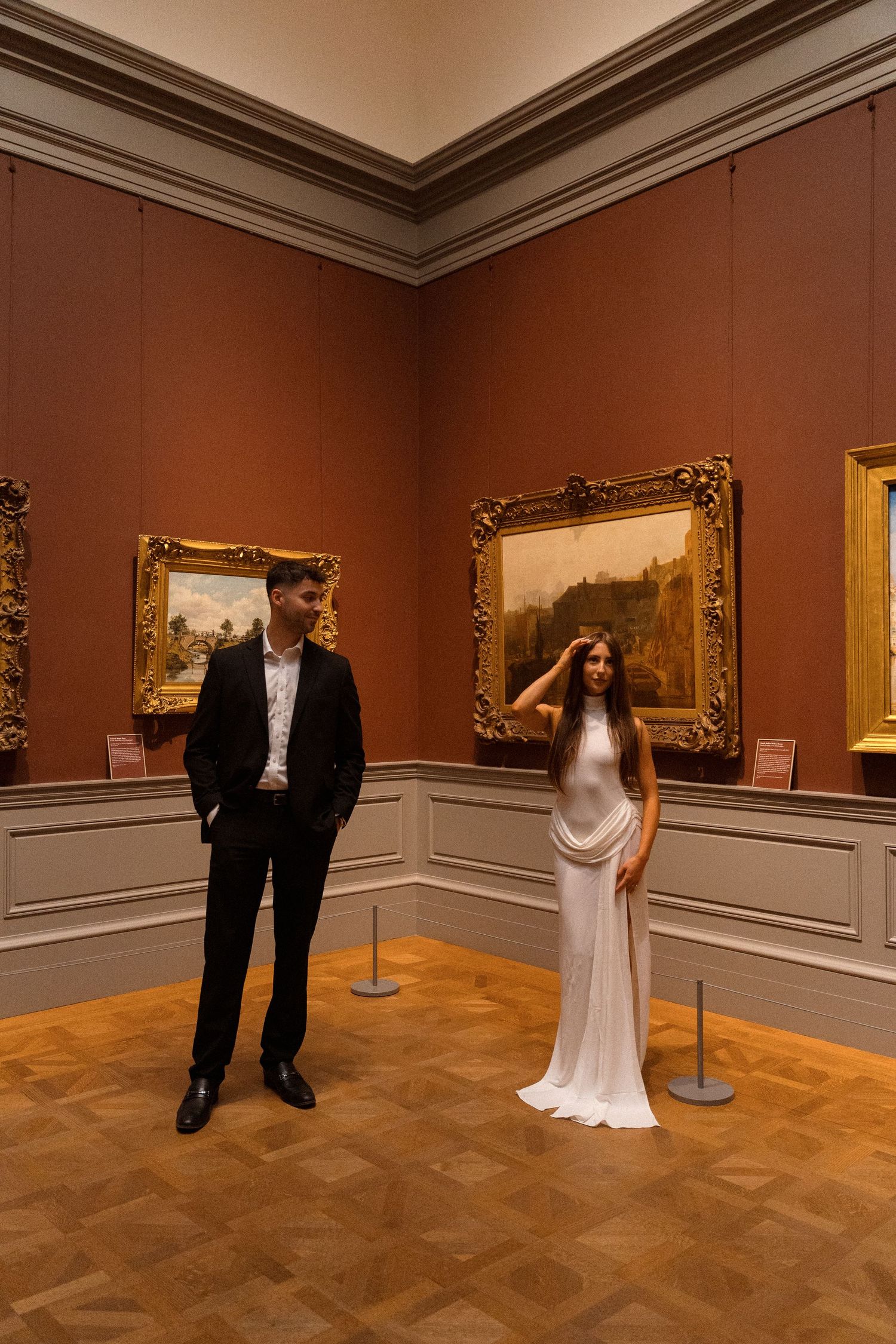 A couple poses playfully in a museum gallery with ornate gold-framed paintings on terracotta walls.
