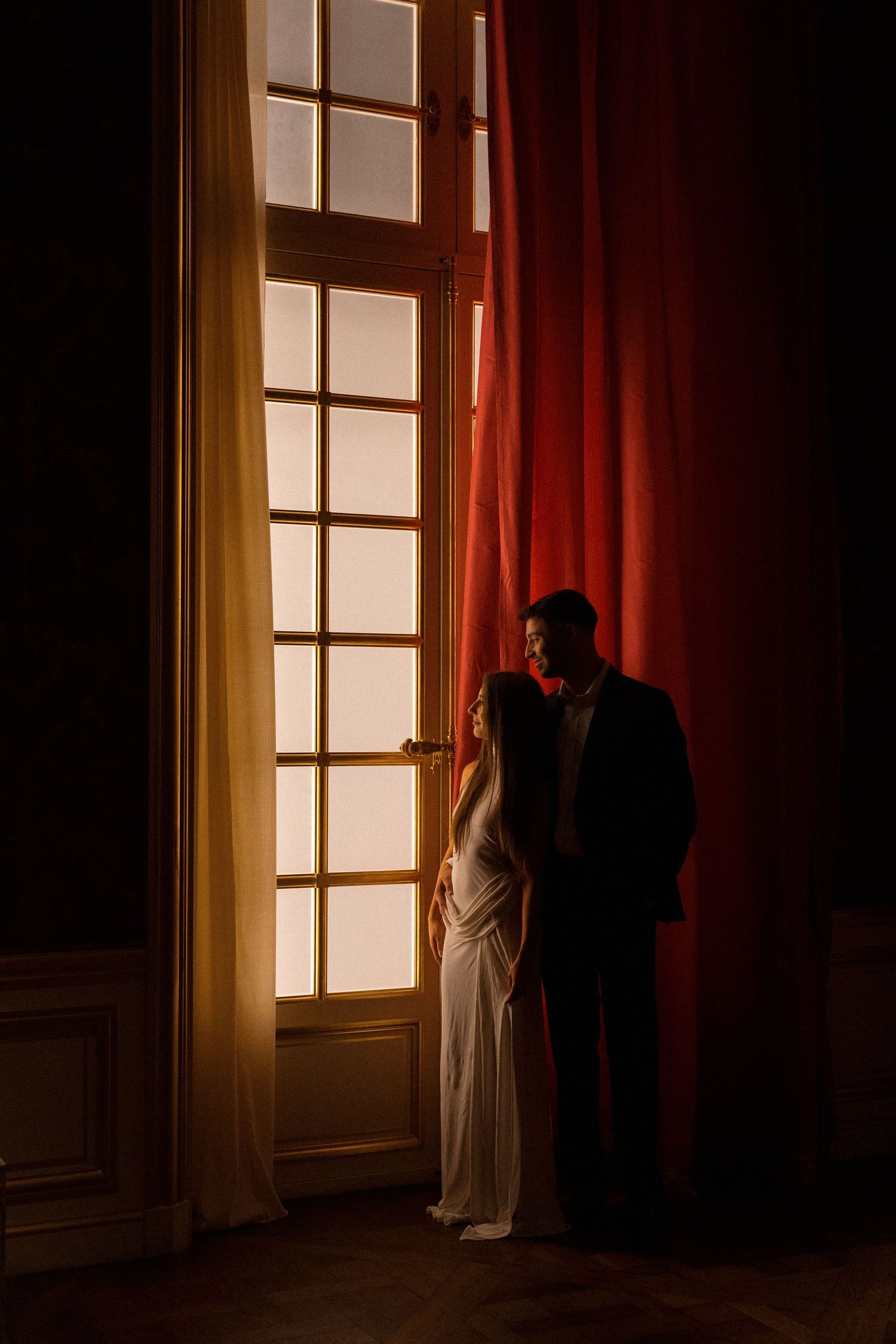 A couple stands in silhouette against a bright window with red curtains creating a dramatic mood.