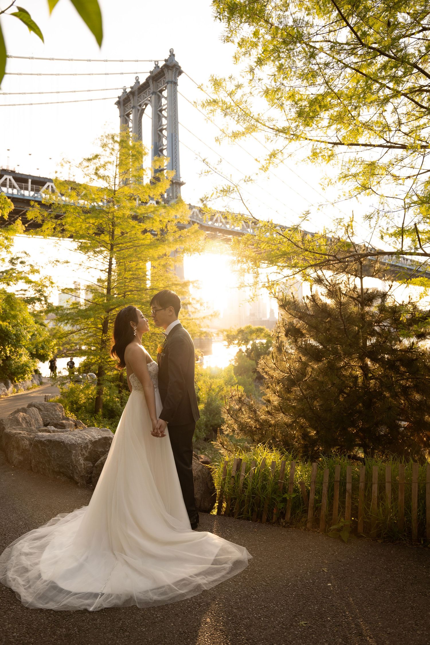 A couple in wedding attire poses against a sunset backdrop with trees and architecture in the background.
