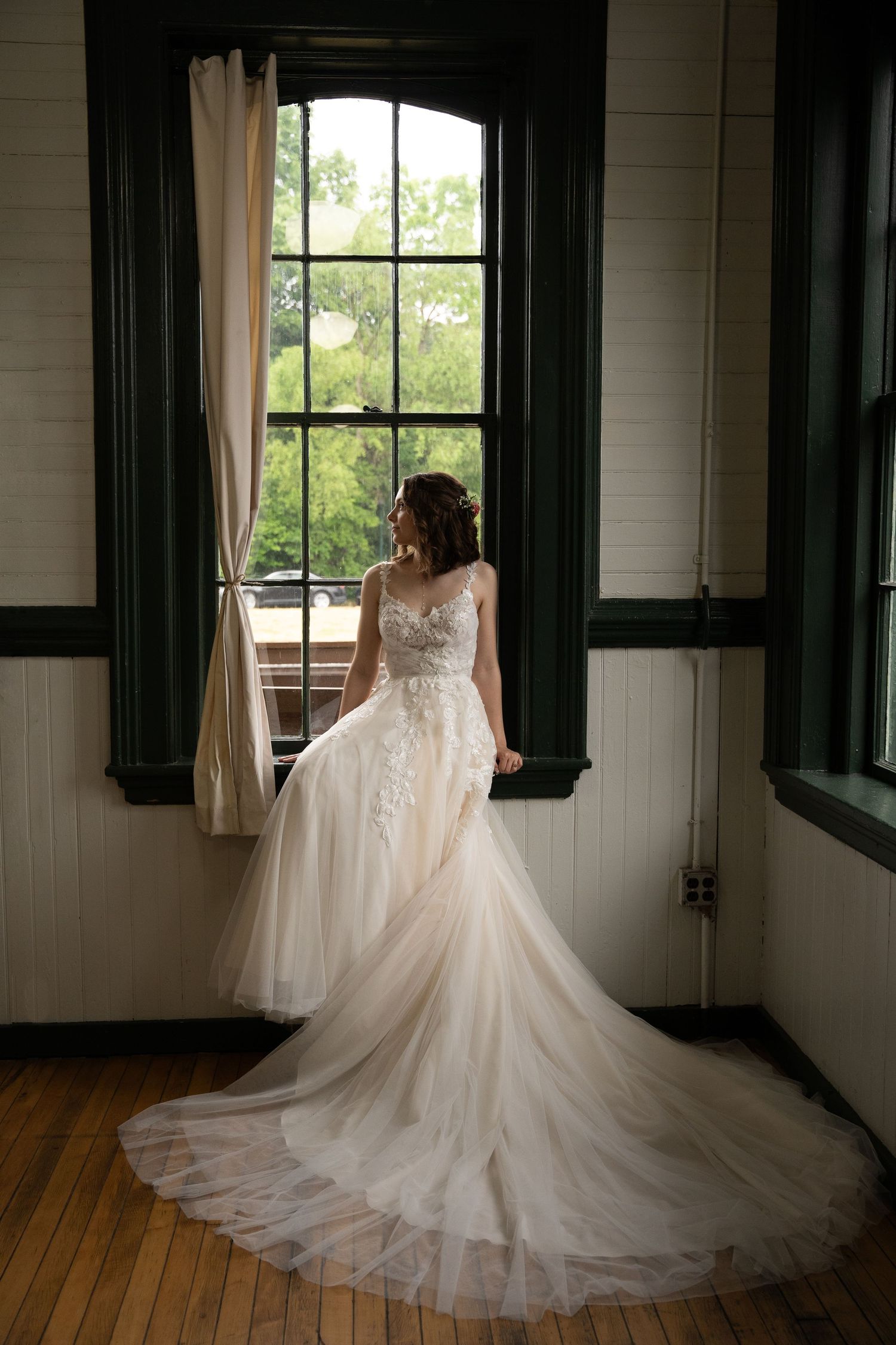 A stunning white wedding gown with a dramatic cathedral train is displayed in a sunlit room with wooden floors.