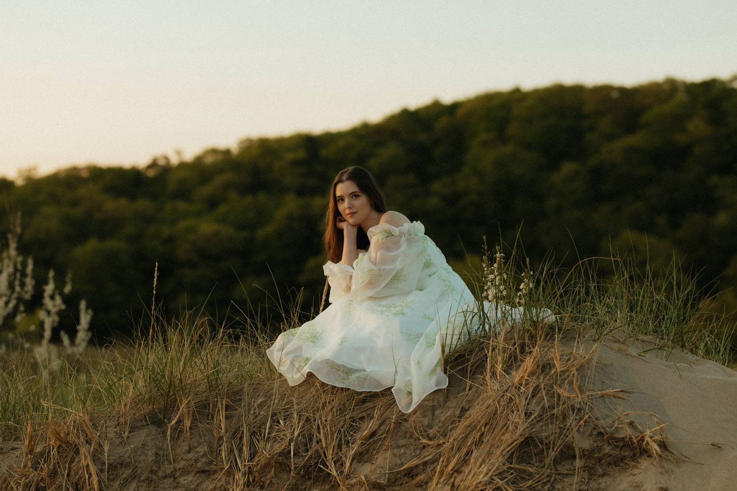 A woman in a flowing white dress sits gracefully on a hillside at sunset.