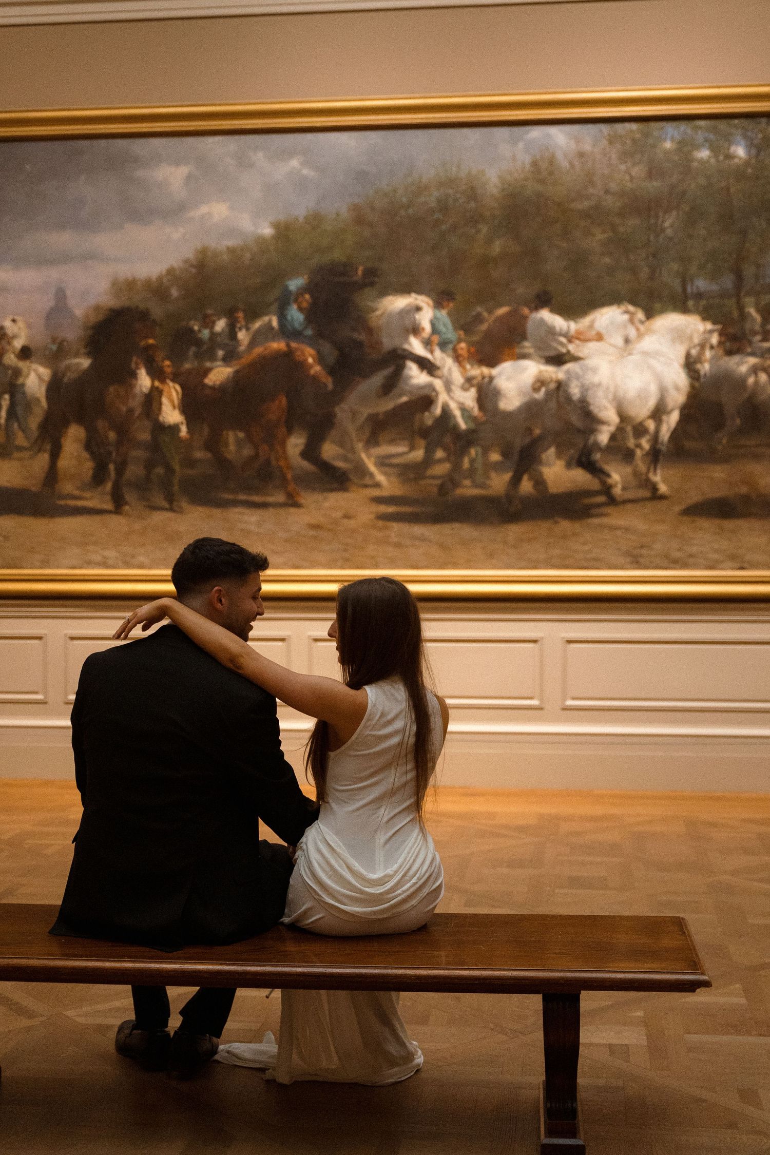 A couple sits on a bench viewing a dramatic horse painting in an art museum.