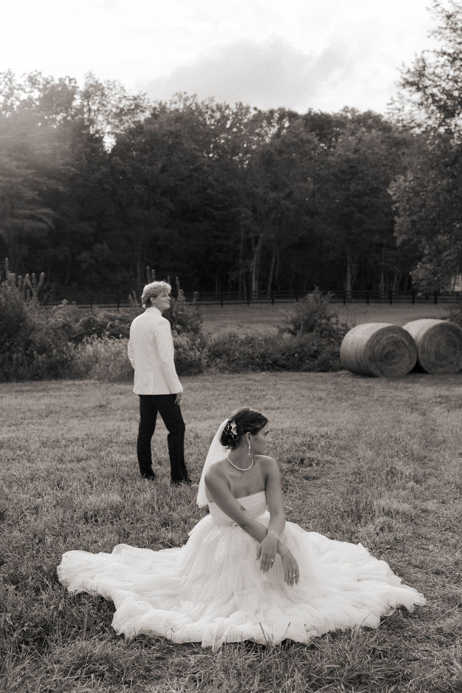 A couple in wedding attire sits together in a peaceful meadow surrounded by trees.