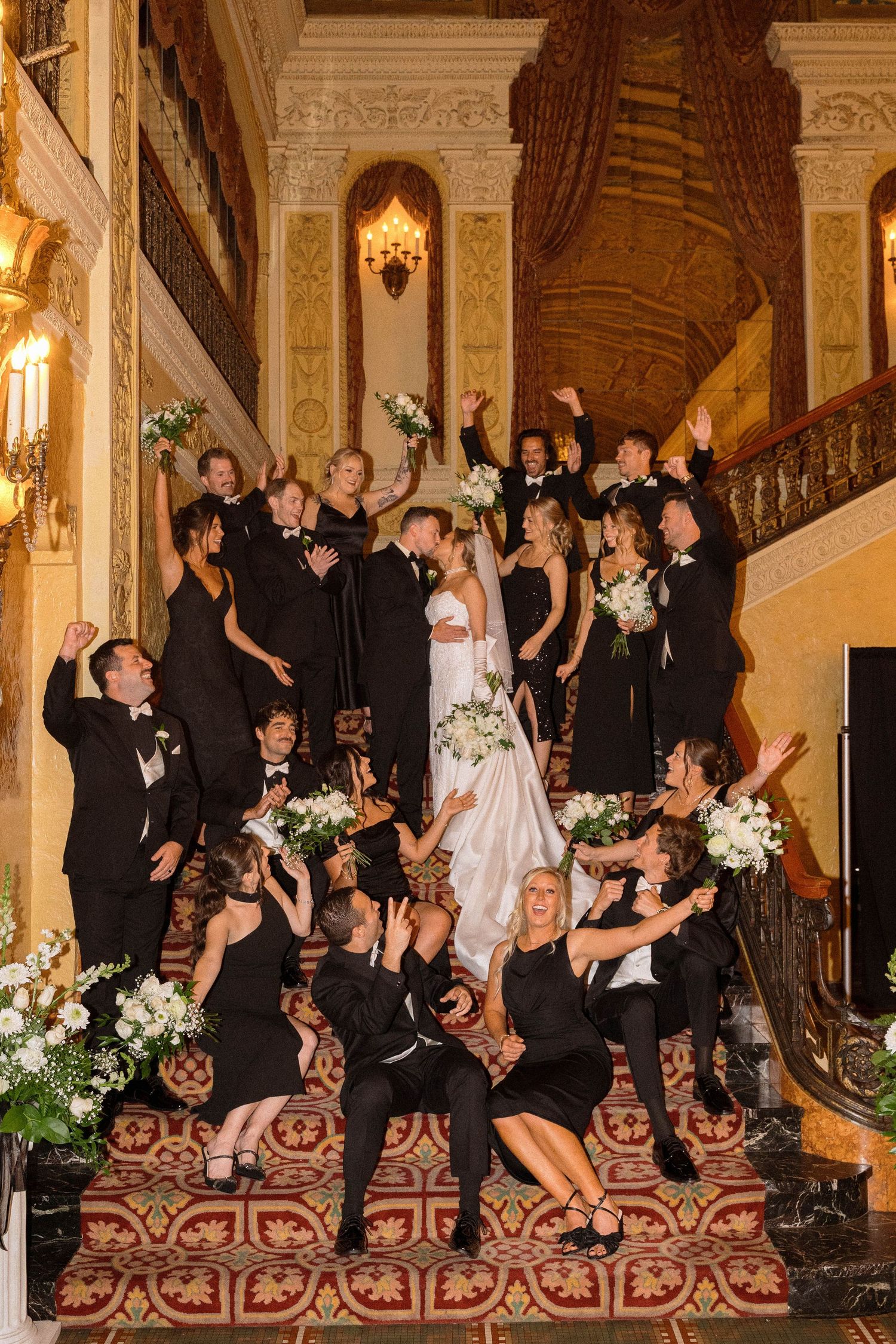 Large wedding party group photo on ornate staircase with guests in formal black attire.