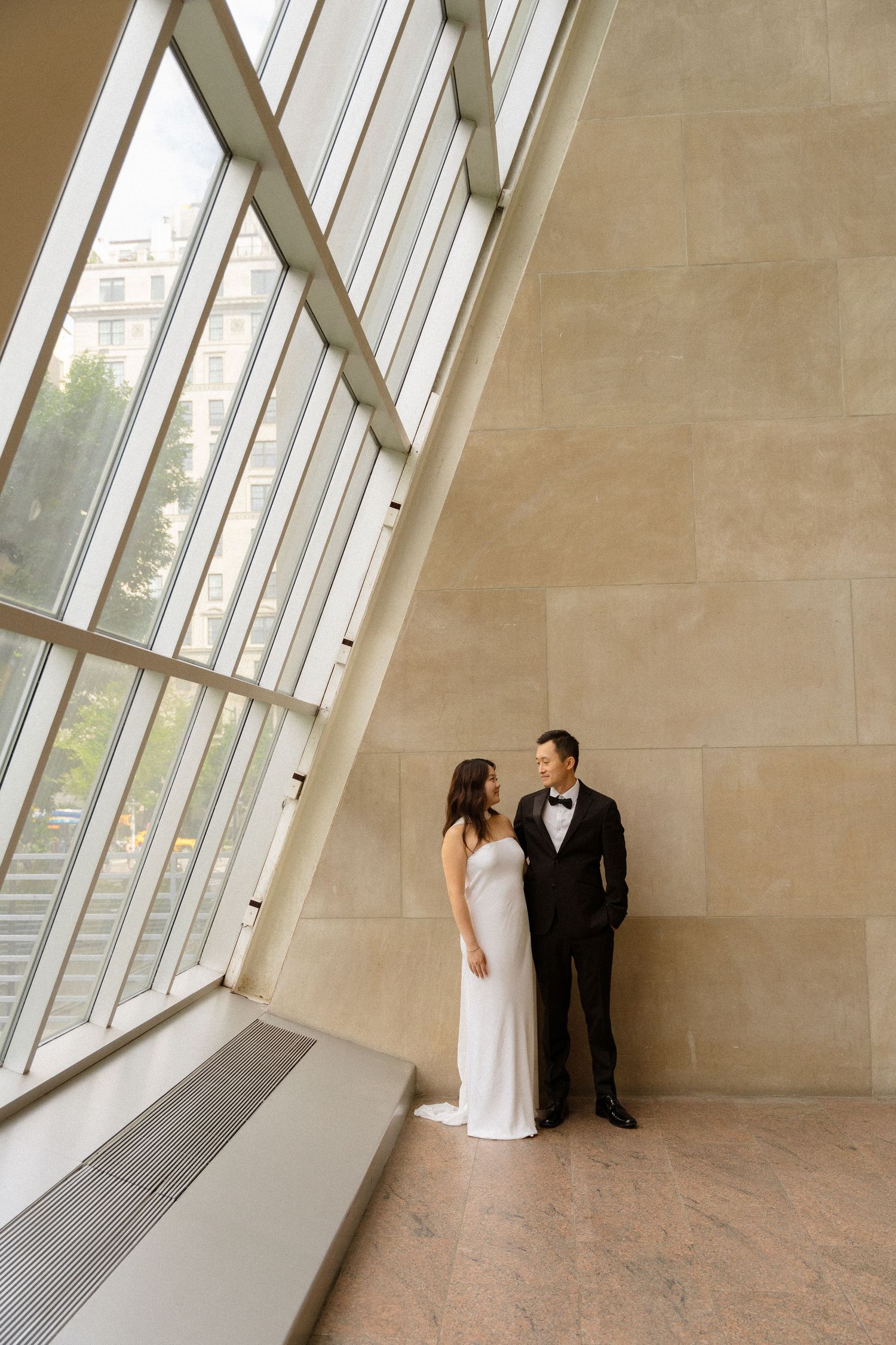 A couple in formal attire poses near angled windows in a modern architectural space.