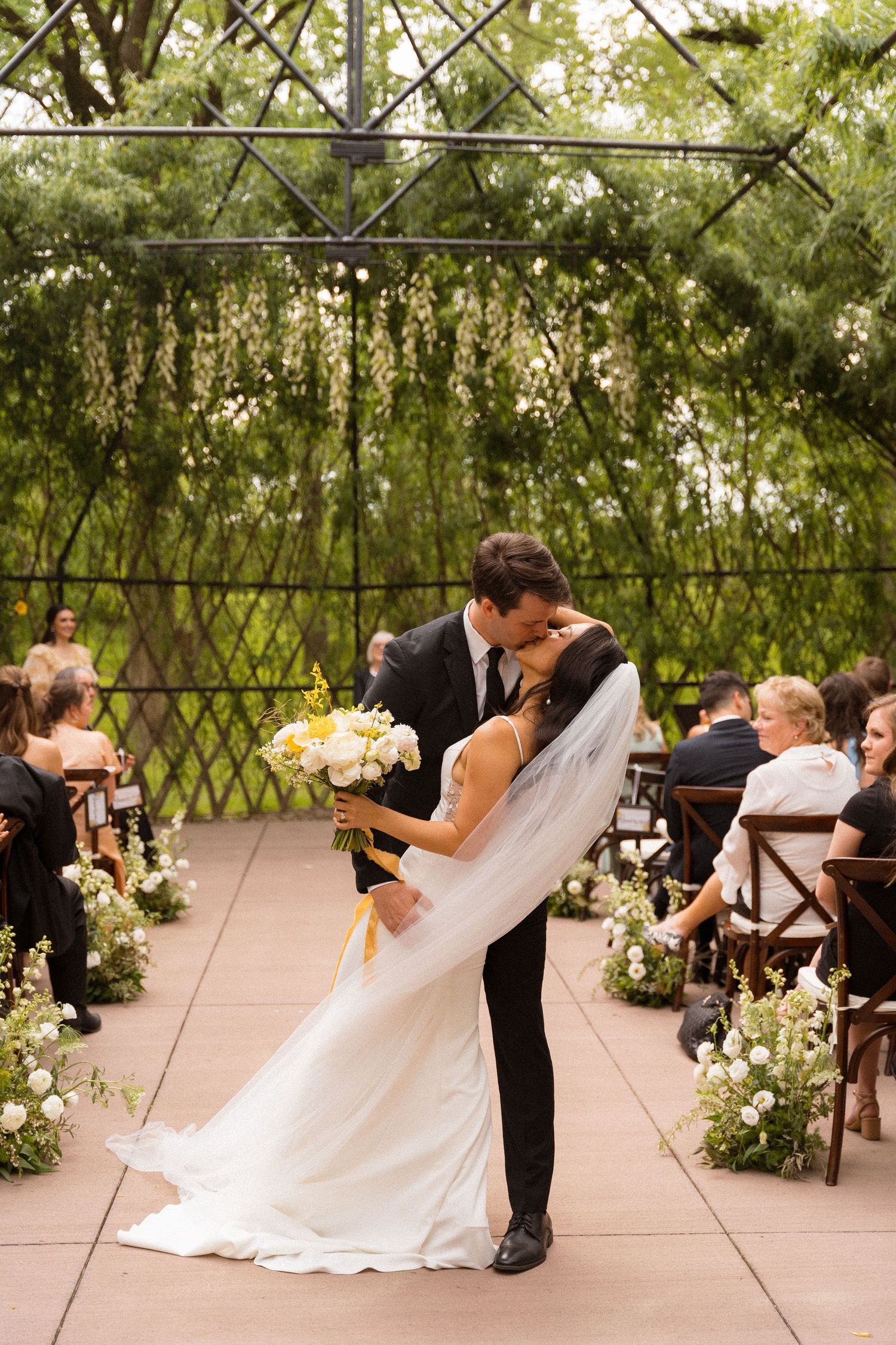 A wedding ceremony taking place under a lush garden trellis with white floral decorations.