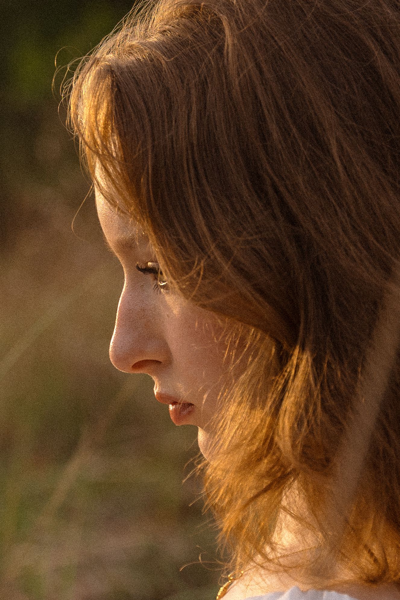 Close-up profile portrait with soft lighting highlighting brown hair against a blurred natural background.
