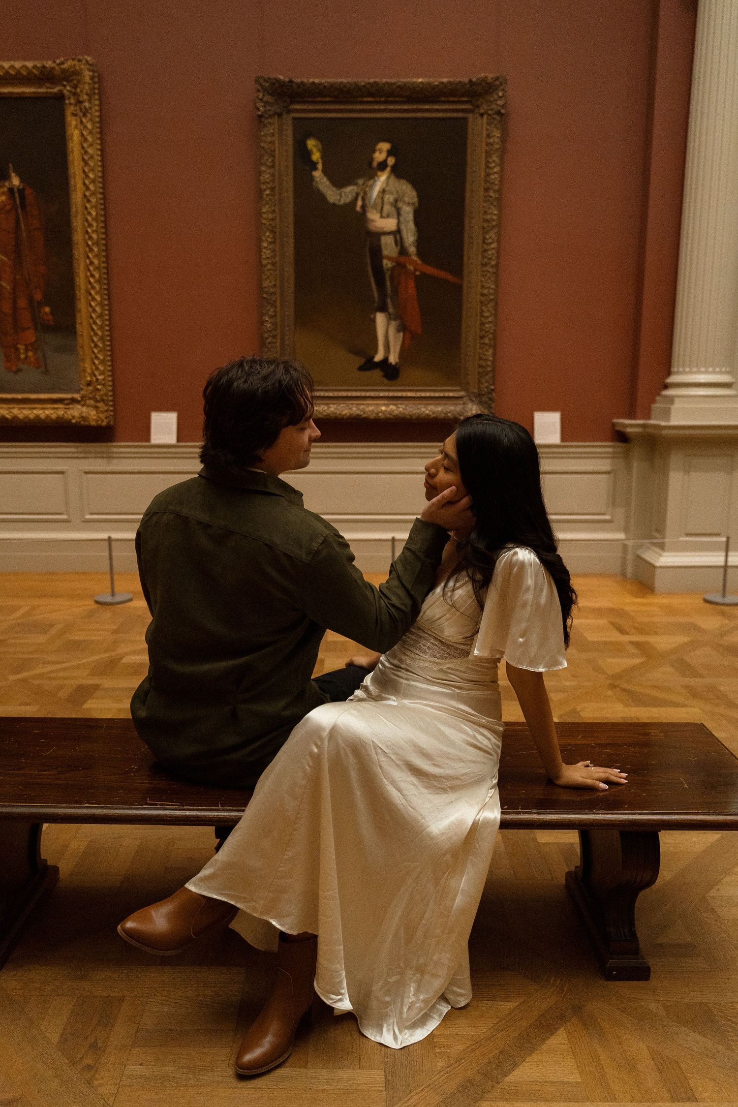 A couple sits together viewing artwork in an elegant museum gallery with wooden floors and ornate frames on the walls.