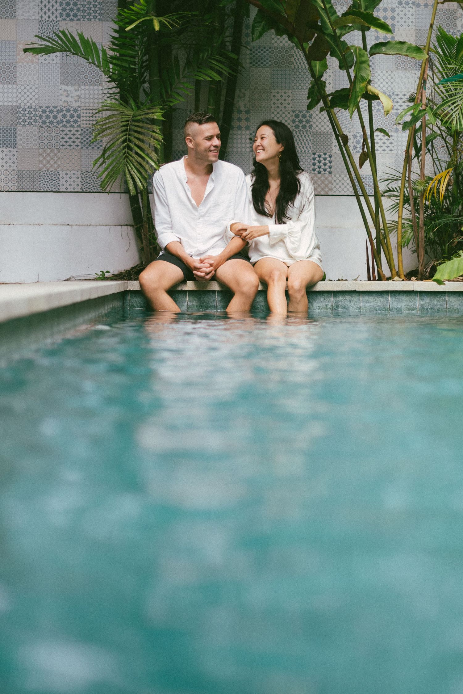 Couple in white shirts sitting poolside with feet in water, surrounded by tropical greenery.