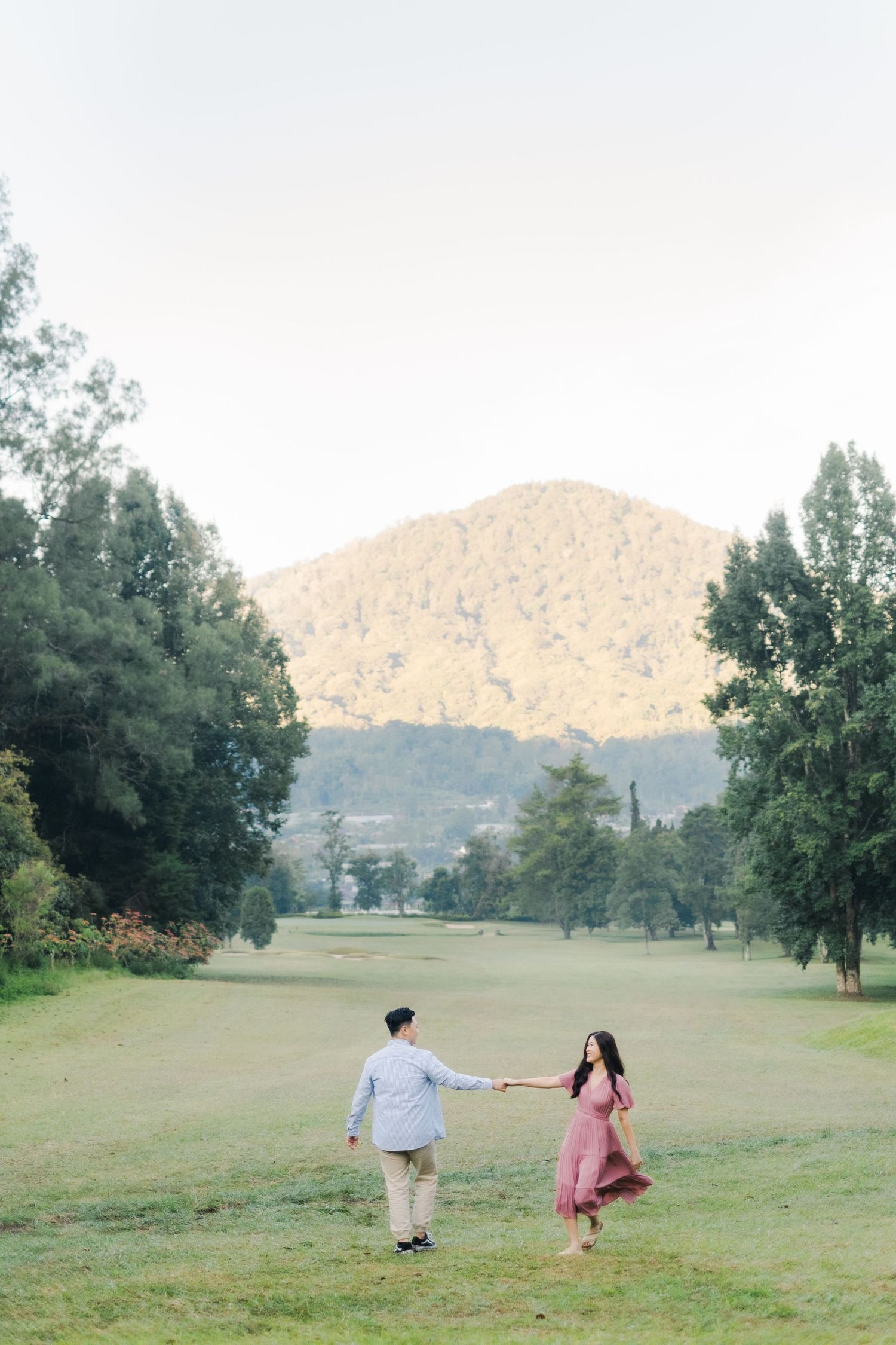 Couple holding hands walking on lush green field with mountain backdrop during golden hour engagement photo session.