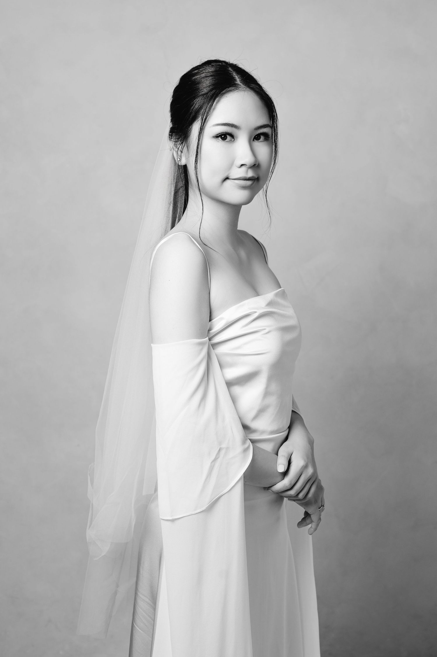 Black and white portrait of a bride in a sleek white gown and veil, posing gracefully against a neutral background.