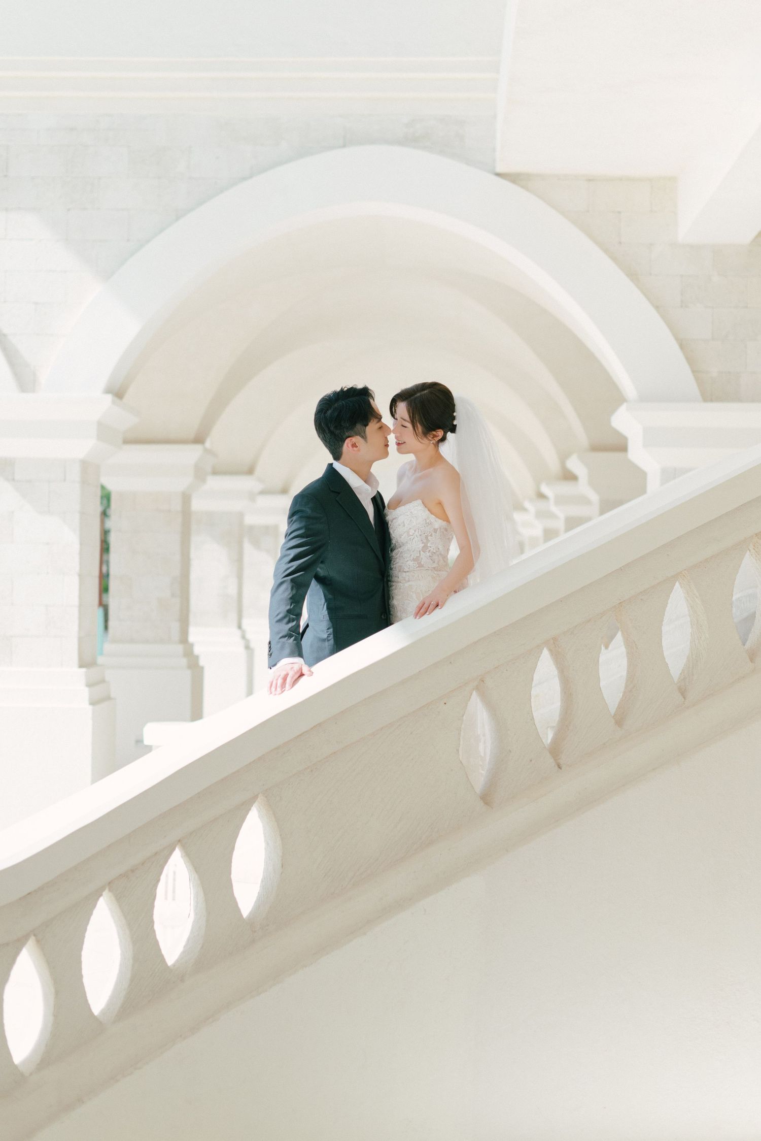 Bride and groom share a kiss on elegant white marble staircase with arched columns in a grand wedding venue.