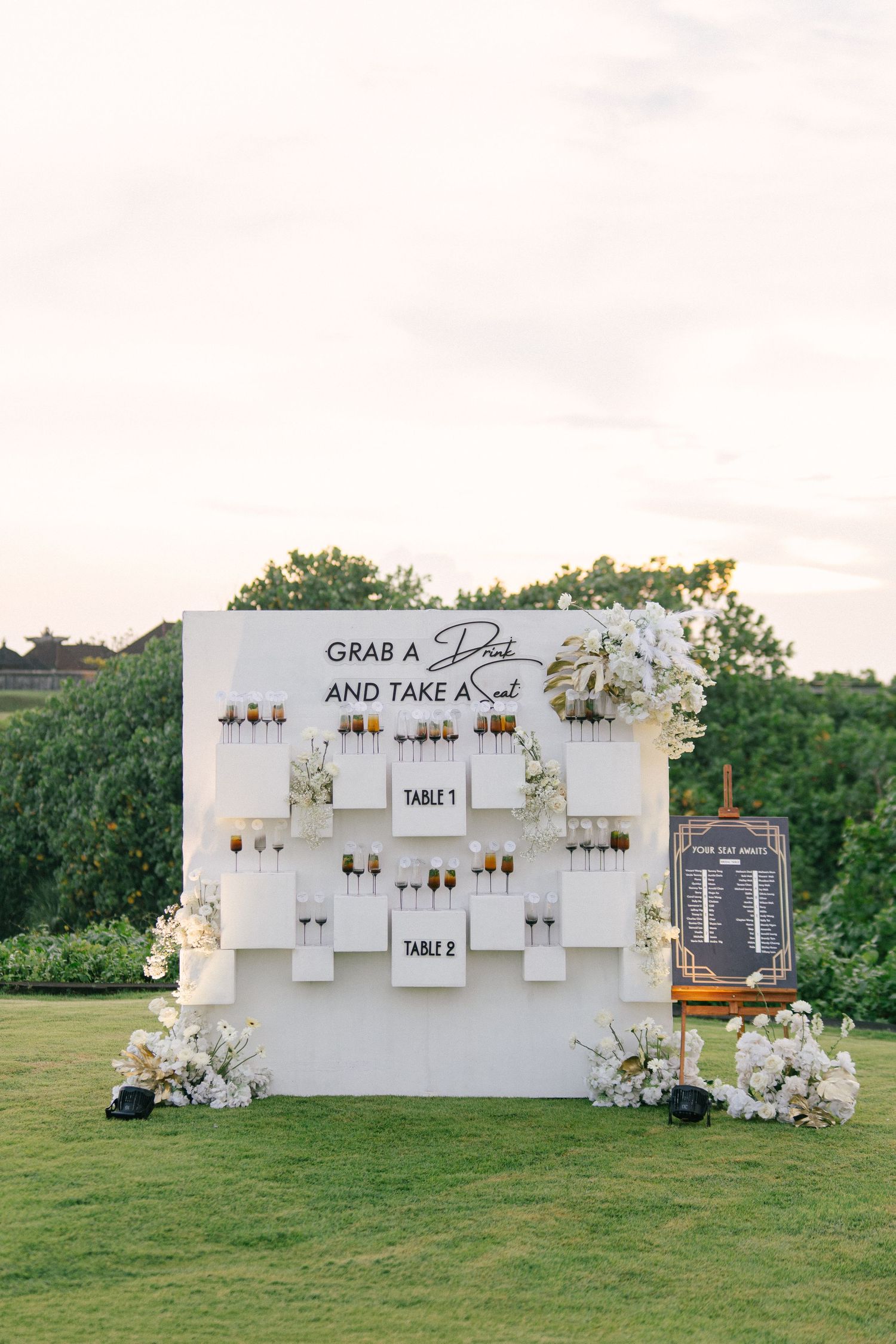 Elegant wedding drink wall with white shelves, champagne flutes, floral decor, and a grab a drink and take a seat sign.