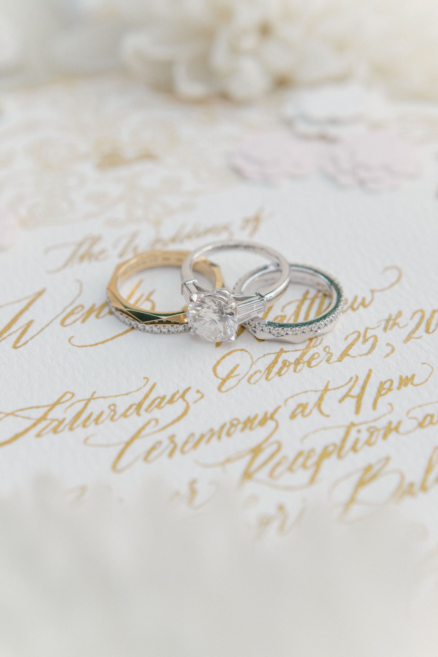 Wedding rings and diamond engagement ring displayed on elegant gold calligraphy invitation for Saturday October 25th ceremony.
