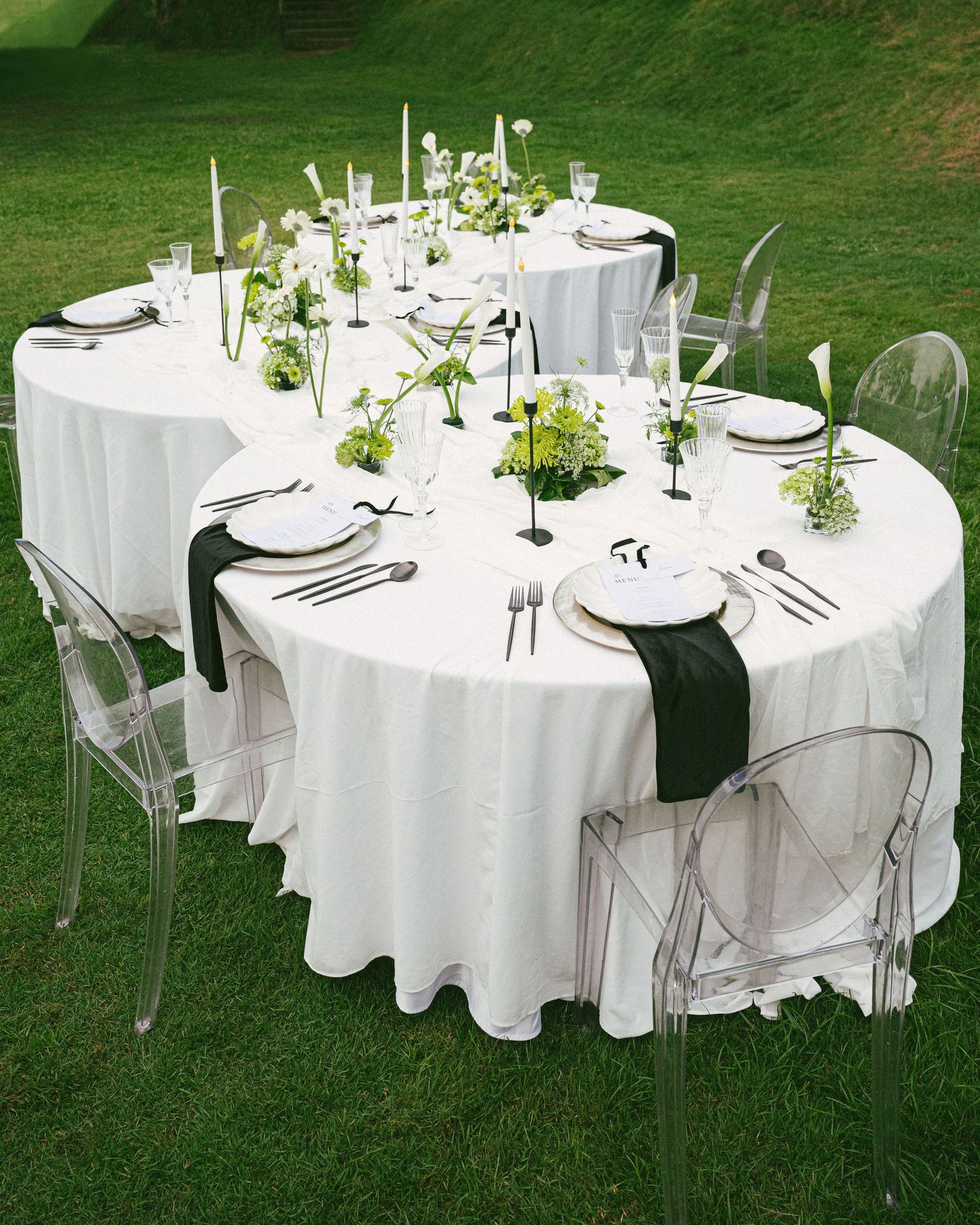 Elegant outdoor wedding reception with white round tables, ghost chairs, green floral centerpieces, and black table runners on grass.