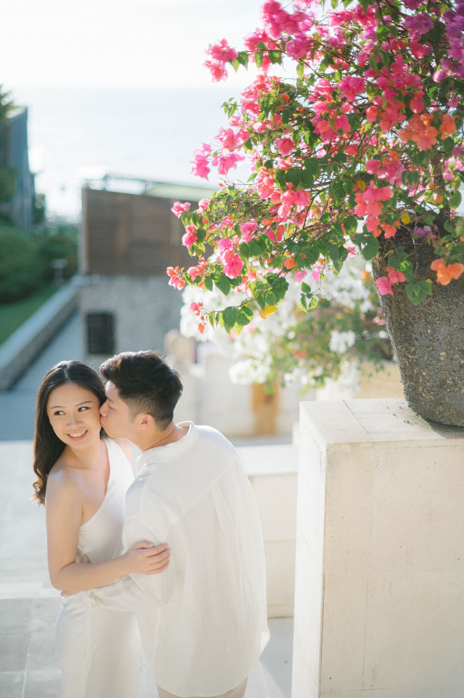 Couple in white embracing near vibrant pink bougainvillea flowers during romantic outdoor photo session.