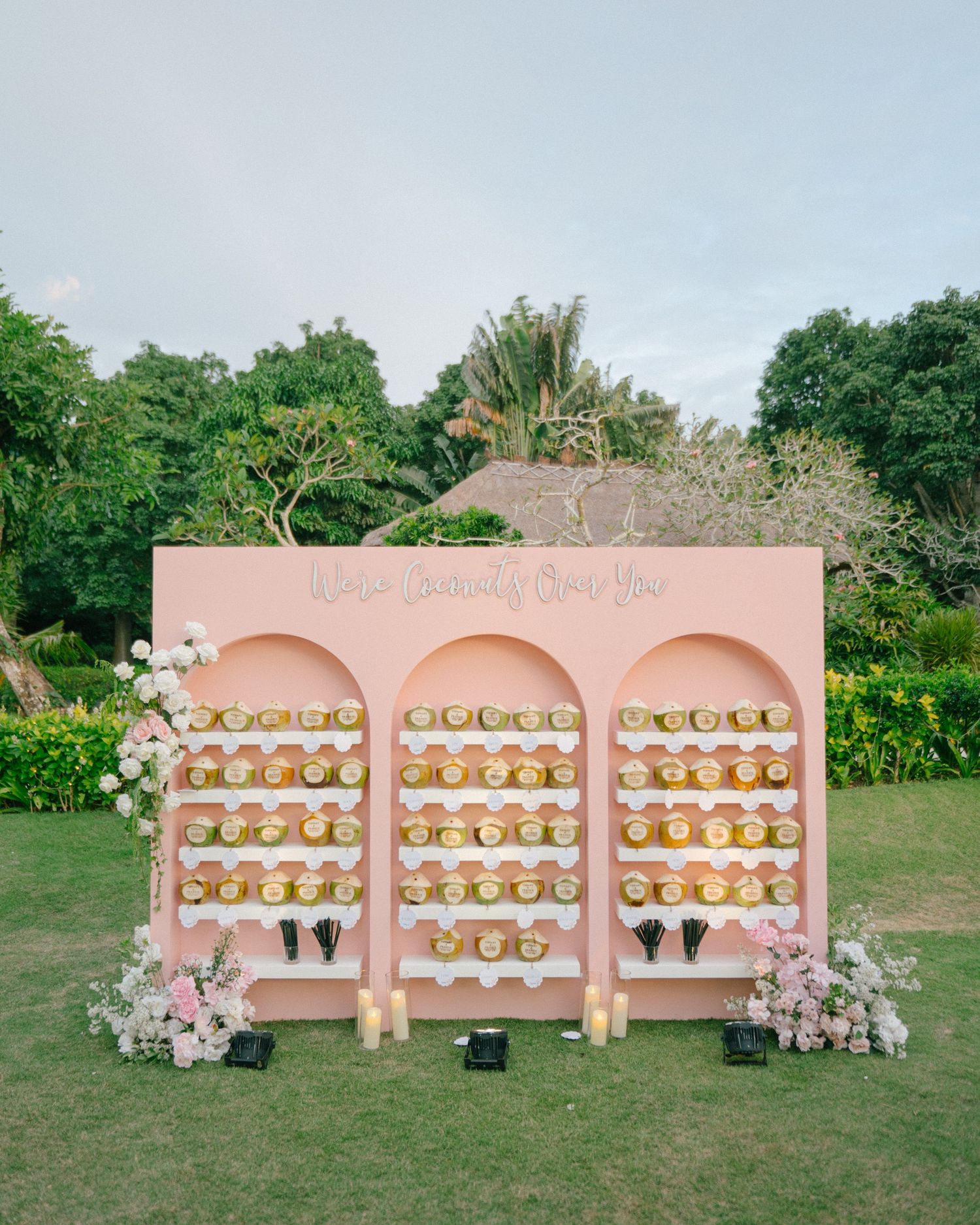 Pink wedding seating chart display with floral arrangements and golden escort cards on outdoor lawn at elegant garden venue.