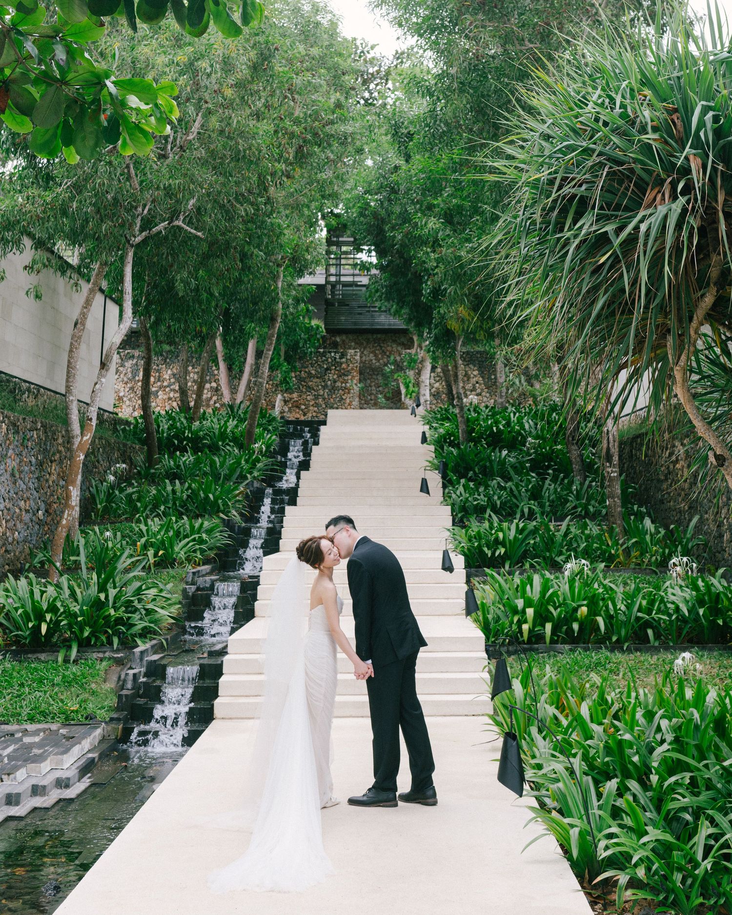 Bride and groom share a romantic kiss on elegant white stairs surrounded by lush tropical gardens at a destination wedding.