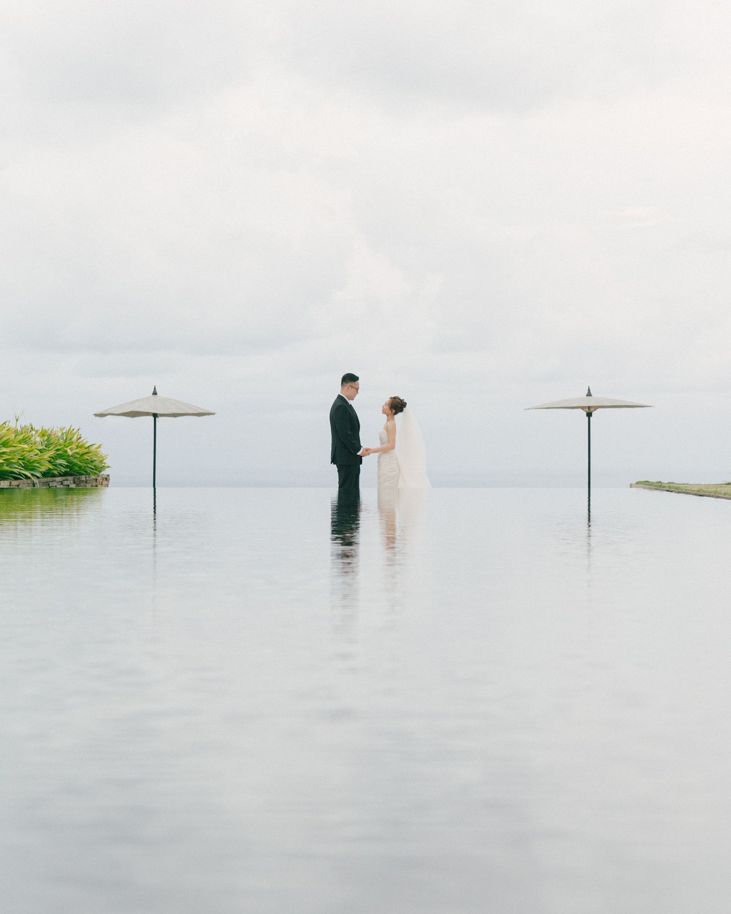 Bride and groom hold hands at infinity pool edge with ocean backdrop and white umbrellas on overcast wedding day.