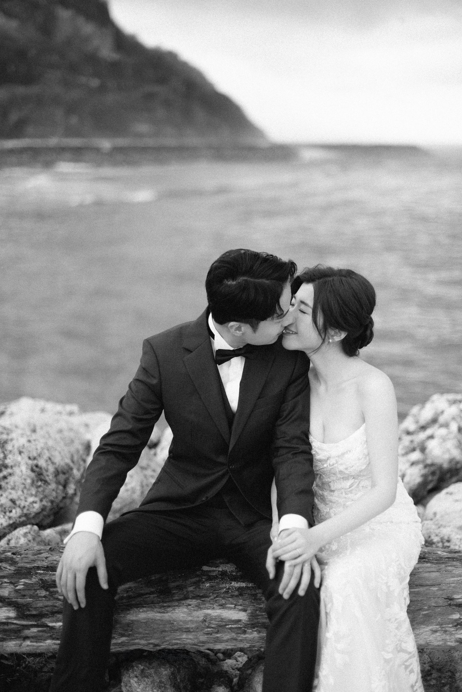 Black and white photo of a couple in wedding attire sharing a kiss on rocks by the ocean.