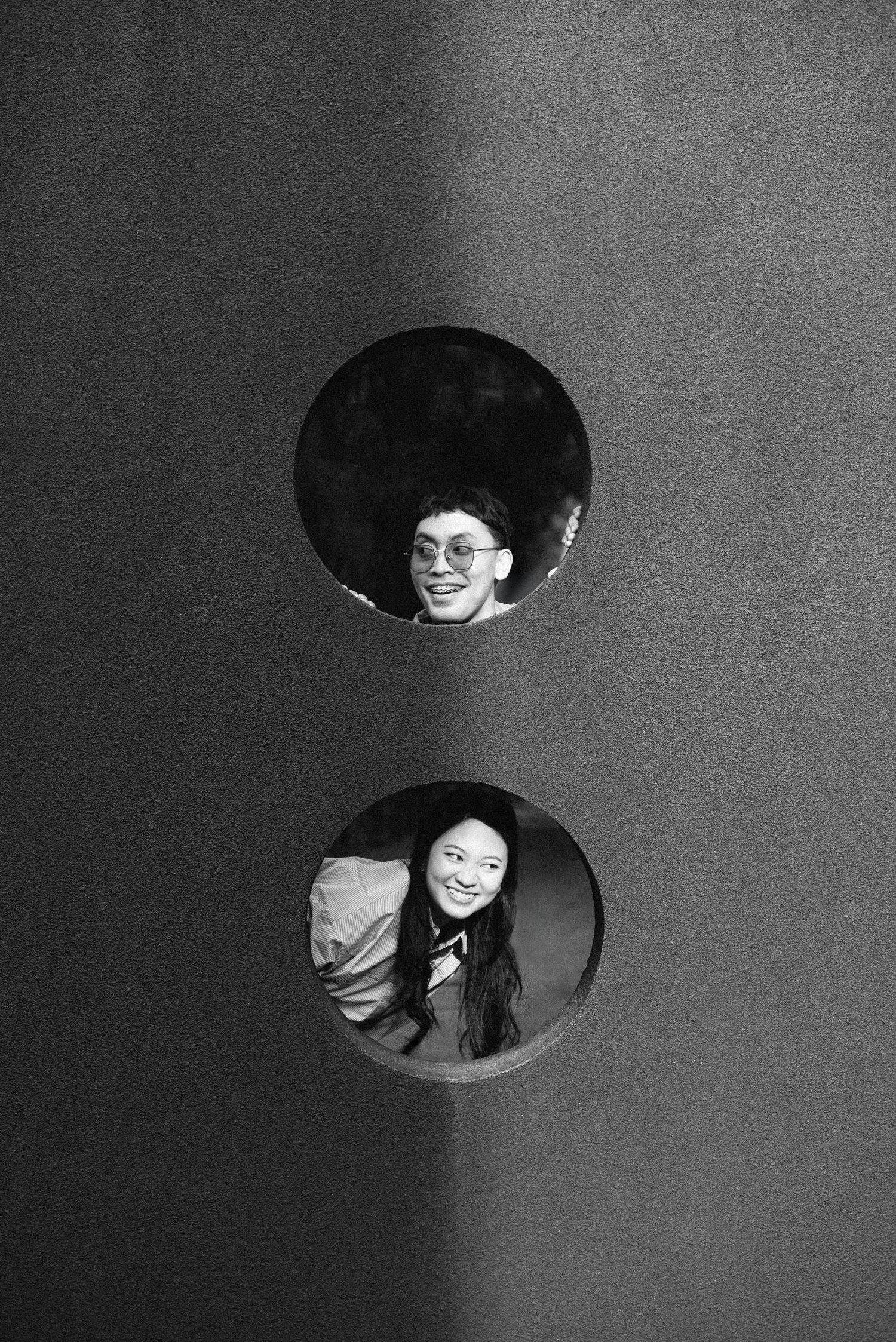 Black and white photo of two people smiling through circular holes in a wall.