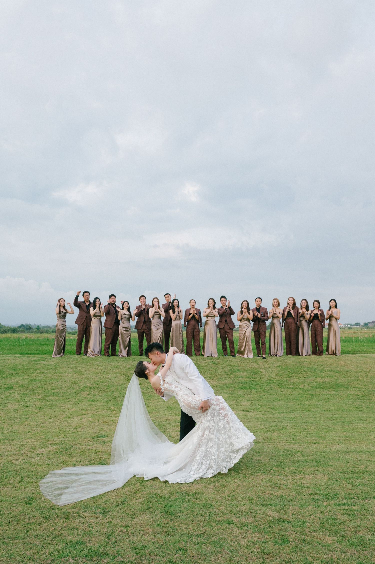Bride and groom share a romantic dip kiss on green lawn with wedding party in brown and taupe attire behind them.