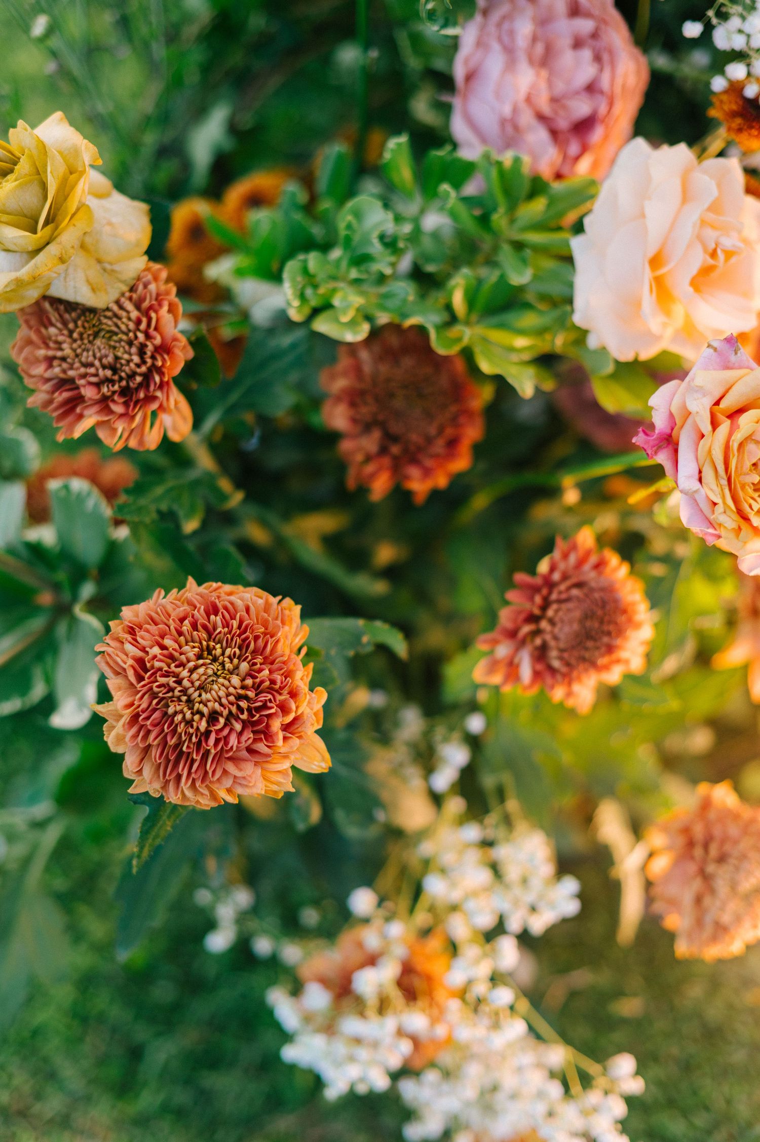 Lush floral arrangement featuring orange chrysanthemums, pink and yellow roses, and green foliage in warm autumn tones.