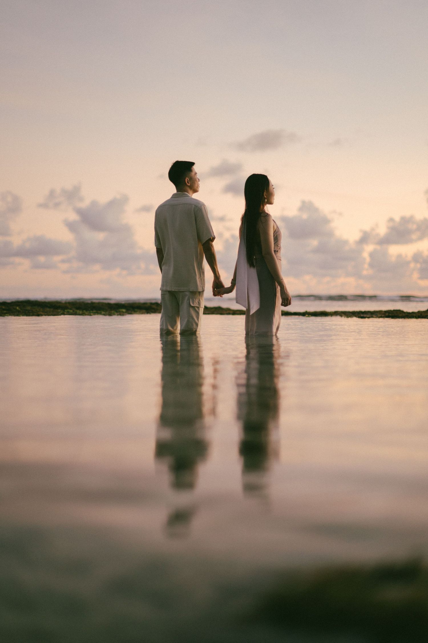 A couple holds hands standing in shallow ocean water at sunset, their reflections visible on the calm surface.